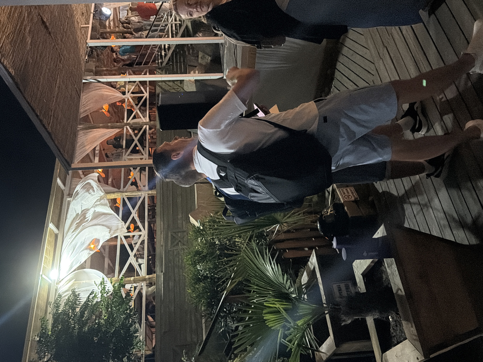 The image depicts an outdoor nighttime scene at what appears to be a venue in Sirius, Russia. The setting is a wooden deck with several people present, some standing and others on a set of stairs leading to a raised area. The raised area is covered by a thatched roof and draped with white fabric, suggesting it might be an outdoor lounge or event space. Orange lanterns are visible, providing some illumination.
In the foreground, a man wearing a white polo shirt and light blue shorts, with a black backpack, is facing left and appears to be talking or gesturing. To his right, a woman in a dark jacket is looking towards him. Another person's legs are visible in the lower right corner.
Behind the foreground figures, there are potted plants, including a palm tree. Further back, on the elevated level, groups of people are scattered, some standing and conversing, others ascending or descending the stairs. A large black speaker is positioned on the deck near the man in the white shirt. The overall ambiance suggests a casual social gathering or entertainment event at night.