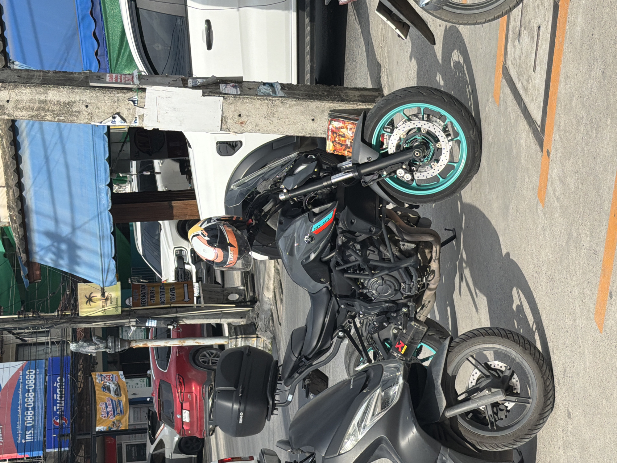 This image depicts a street scene in Baan Mae Nam, Thailand. In the foreground, a dark gray motorcycle is parked, featuring a black luggage box on the rear seat and distinctive turquoise accents on its front wheel rim. To the left of this motorcycle, the front portion of another dark gray scooter is visible, with the "HONDA" logo discernible on its front.
Behind the motorcycles, a white pickup truck is parked, with its front doors and side visible. A red SUV is parked to the left of the pickup truck. In the background, commercial buildings line the street, adorned with various signs and awnings. One sign, featuring a palm tree and the text "SAMUI," is prominent. A concrete utility pole stands near the center of the frame, covered in various flyers and advertisements, one of which appears to be a flyer for tours and travel. The ground is paved with concrete, marked with orange lines indicating parking spaces. The lighting suggests it is daytime, with shadows cast on the ground.