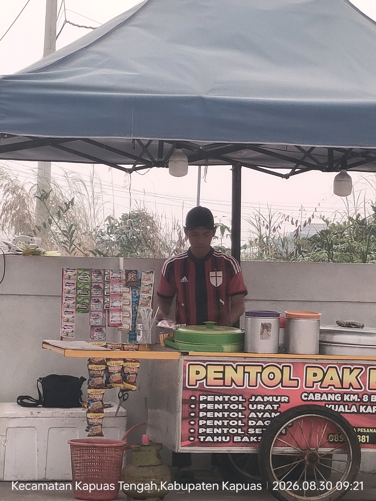 A young man wearing a red and black striped shirt with white stripes on the sleeves and a black cap is standing behind a food cart. The cart has a red banner with yellow text that reads "PENTOL PAK B". Below the banner, a list of pentol types is visible, including "PENTOL JAMUR", "PENTOL URAT", "PENTOL AYAM", "PENTOL DAG", "PENTOL SE", and "TAHU BAK". To the right of the banner, text reads "CABANG KM. 8 B KUALA KAPI". A bicycle wheel is attached to the cart. The cart is filled with various items, including containers, drinks, and snacks displayed on shelves. In front of the cart, there is a red basket and a gas cylinder. The background shows tall grass and foliage, suggesting an outdoor setting. The image is overlaid with text that reads "Kecamatan Kapuas Tengah, Kabupaten Kapuas | 2026.08.30 09:21".
