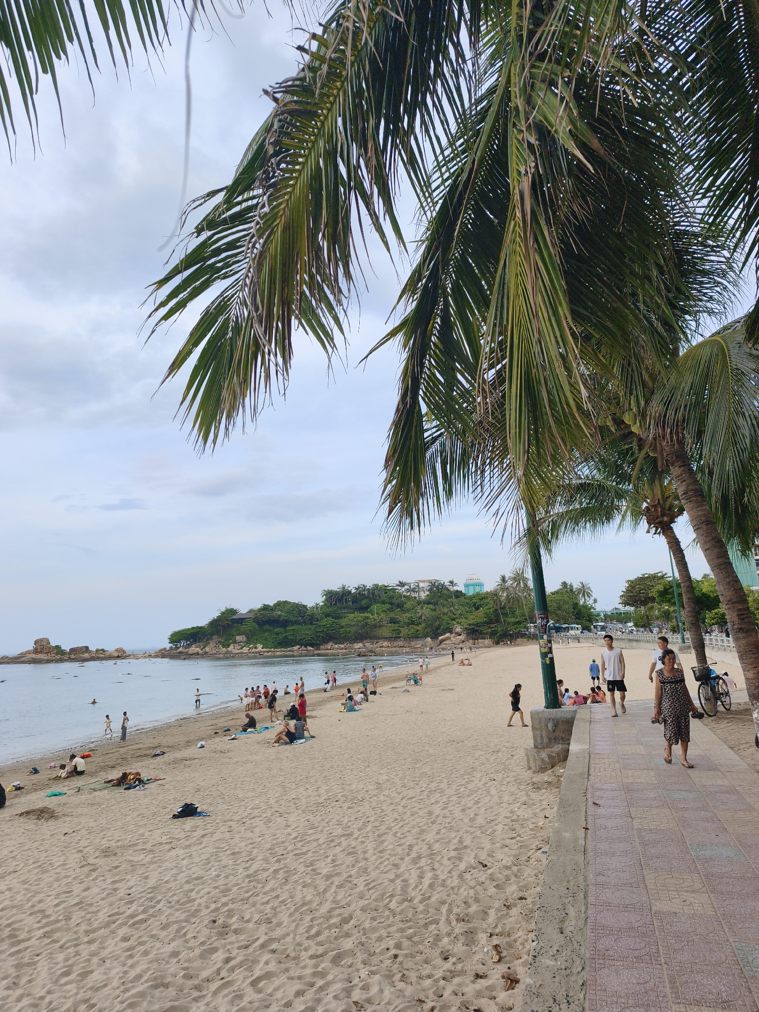 The image shows a beach scene in Nha Trang, Vietnam, likely near the central beach area. The foreground is dominated by the sandy beach with several palm trees casting shadows. There is a paved walkway running alongside the beach, with a few people walking on it. The beach itself is populated with numerous individuals, some sitting on towels, others walking, and a few children playing near the water's edge. The sea is calm, with a distant headland covered in green vegetation and some rocky outcrops visible in the water. In the background, there are buildings and more palm trees, suggesting an urban or semi-urban coastal environment. The sky is overcast with some patches of blue.