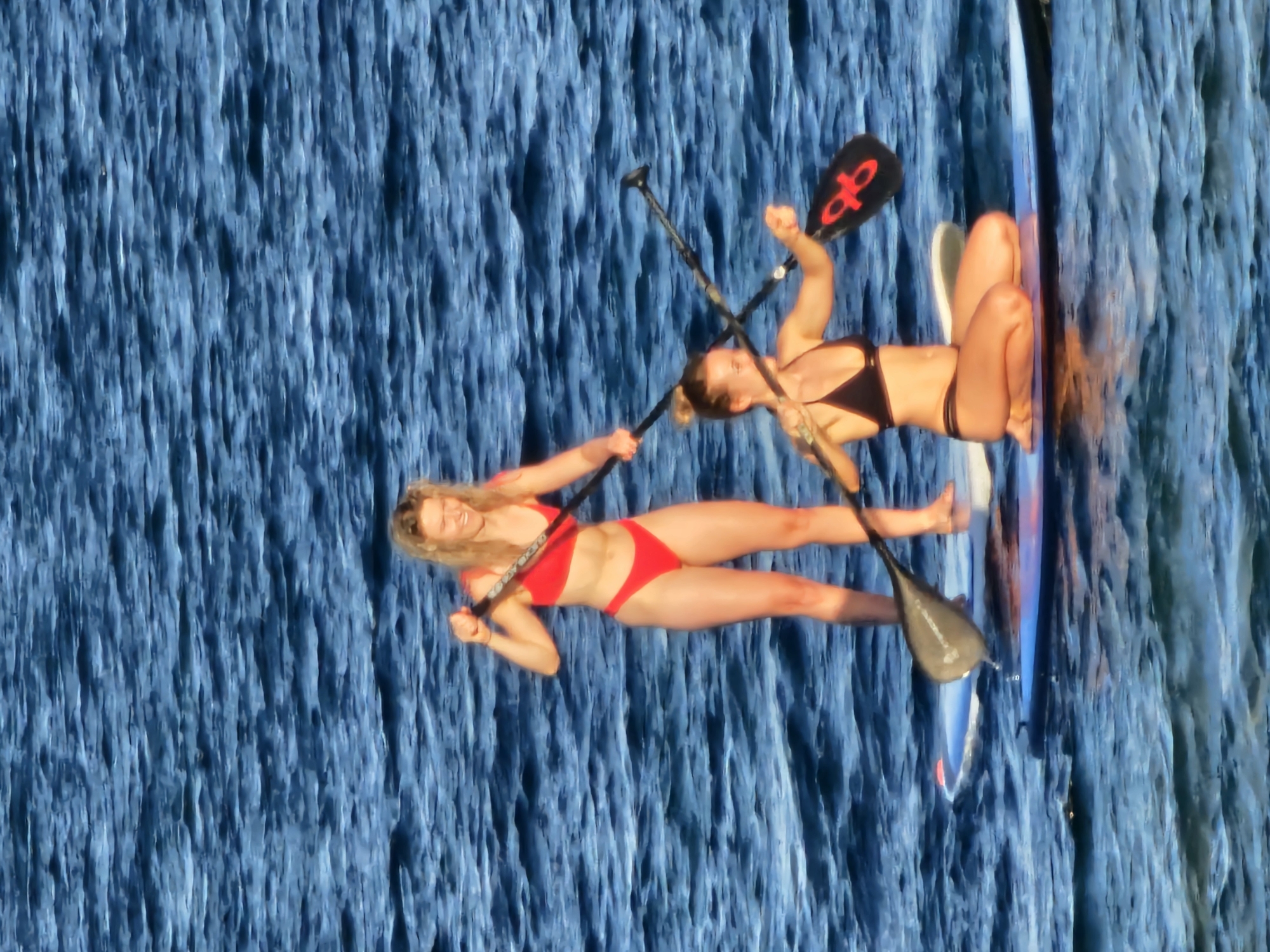 Two women are paddleboarding on the ocean. The woman in the foreground is kneeling on a paddleboard and wearing a black bikini. She is holding a paddle with a red logo. The woman in the background is standing on a paddleboard and wearing a red bikini. She is also holding a paddle. The water is blue and choppy. There are no discernable landmarks or features that would indicate a specific location within Denpasar, Indonesia.