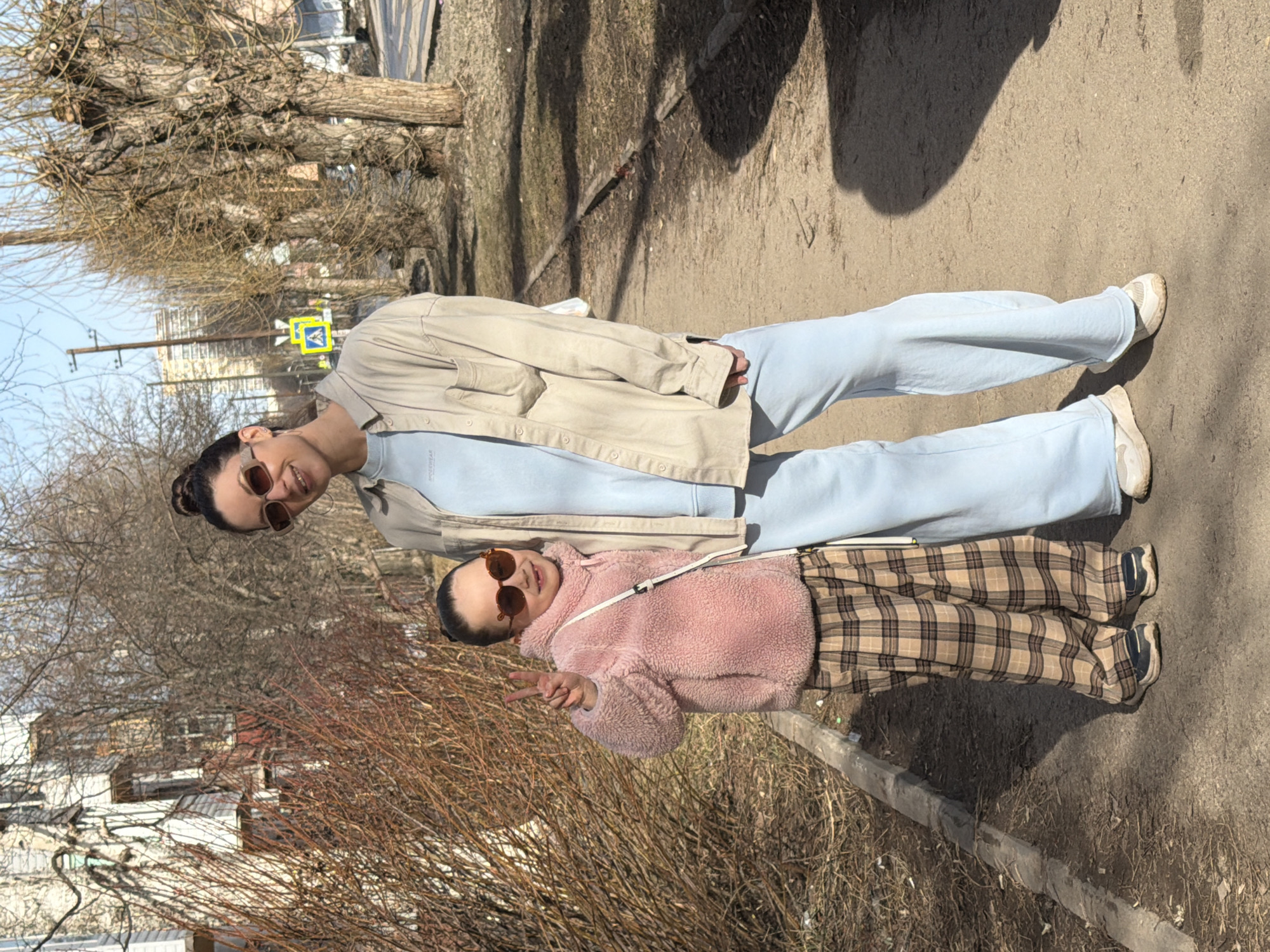 Two women, one older and one younger, stand next to each other on a sidewalk. The older woman wears sunglasses, a light blue sweater, light blue pants, and a tan jacket. The younger woman wears sunglasses, a pink fluffy jacket, and plaid pants. The younger woman holds up her hand in a peace sign. Behind them, a building and bare trees are visible. They are located in Perm, Russia, evidenced by the street sign indicating pedestrian crossings.