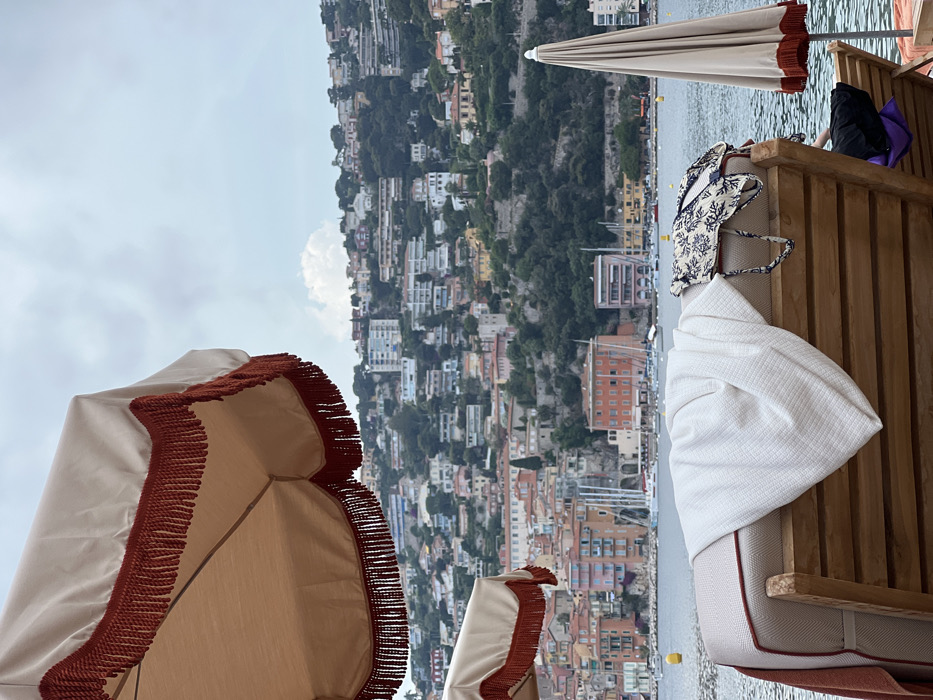 Captured from a low angle, the image showcases beach umbrellas with tasseled edges, casting shade on wooden lounge chairs. A white patterned beach bag rests on one chair beside a folded white towel. In the background, a densely populated hillside town slopes towards the sea. The scene is filled with light, suggesting a sunny day at the beach.