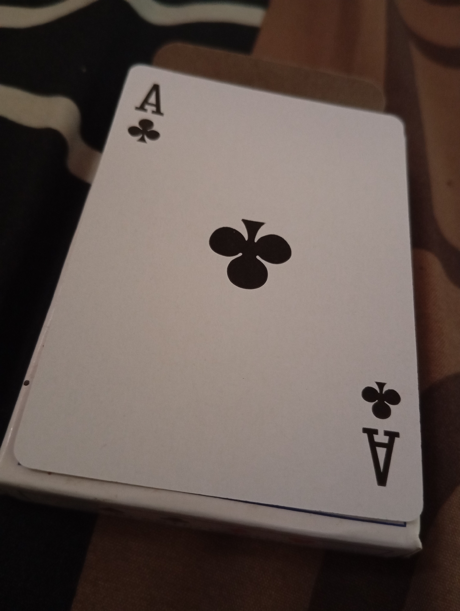 The image shows a close-up, slightly angled view of an Ace of Clubs playing card lying on top of a deck of cards. The card is white with black markings. In the top left corner, there is a black "A" above a black clover symbol. In the center of the card, there is a large black clover symbol. In the bottom right corner, there is a black clover symbol above a black "A". The deck of cards below appears to be white with a blue edge. The background is out of focus and appears to be a patterned surface with black, white, and brown tones. There are no people visible in the image. The lighting is dim, suggesting an indoor setting. There is no information to suggest this image is located in Sudikampiran, Indonesia.