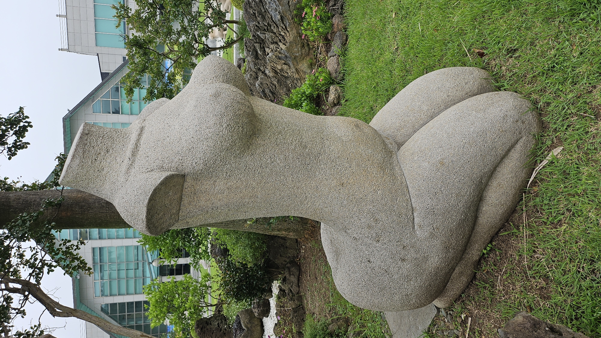 A stone sculpture of a nude female torso and legs is situated on a grassy area in Seogwipo-si, South Korea. The sculpture, made of rough-textured stone, depicts a kneeling woman with her head and arms absent. The torso displays prominent breasts and a defined waist, transitioning into large, rounded thighs and calves. The sculpture rests on a dark, flat stone base.
In the background, a modern building with a distinctive angular roof and large glass windows is visible. Lush green trees and bushes surround the sculpture and the building, suggesting an outdoor park or public space. Rocks and some sparse vegetation are scattered around the base of the sculpture. The sky is overcast, indicating a cloudy day. The overall setting appears to be a well-maintained outdoor area, likely a cultural or artistic space within the city.