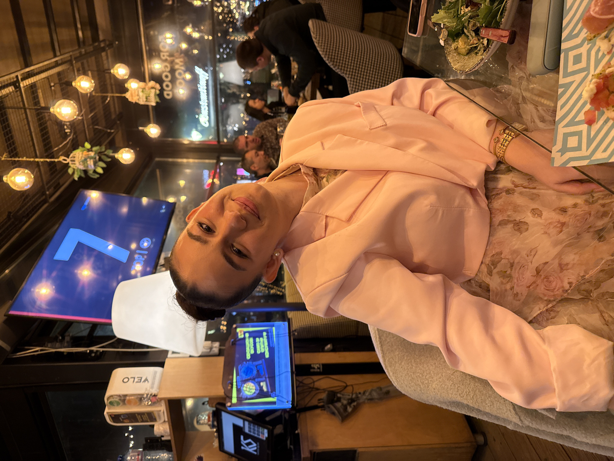 The image shows a woman in a pink blazer at a restaurant, likely in Sofia, Bulgaria, based on a window sign. She's seated at a table with food and drinks, surrounded by other patrons. A TV screen in the background displays a large number "7" and the word "Idea". The scene suggests an evening or night setting, given the lighting and the window view of the city lights.