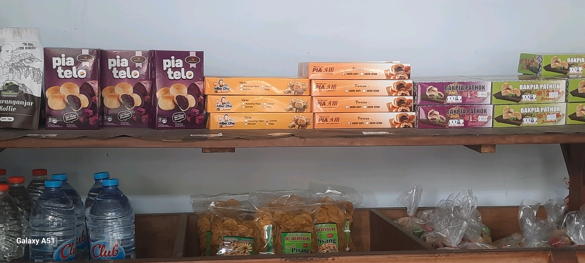 The image shows a retail shelf in what appears to be a shop in Wlingi, Indonesia. The shelf is stocked with various packaged food items, primarily boxes of "Pia Telo" and "Bakpia Pathok," which are traditional Indonesian pastries.
On the top shelf, from left to right, there is a bag of "Karanganyar Koffie" and several boxes of "Pia Telo" with a purple and white design, depicting pastries and purple sweet potato. Next to these are stacks of orange boxes labeled "Mbo'Dhe" and "Pia Am," showcasing different pastry varieties like "Kacang Hijau," "Coklat," "Durian," and "Keju." Further to the right, there are green and purple boxes of "Bakpia Pathok" from Jogja, Indonesia, with text indicating "100% Barokah."
The lower shelf contains several plastic bottles of water, some with red caps and others with blue caps. The brand "Club" is visible on some of the bottles. Also on the lower shelf are transparent plastic bags filled with what appear to be dried banana chips or crackers, with labels indicating "Bu Murvanti" and "Pisang." In the background, other assorted packaged goods in plastic bags are visible. The overall environment suggests a small local store or market stall.