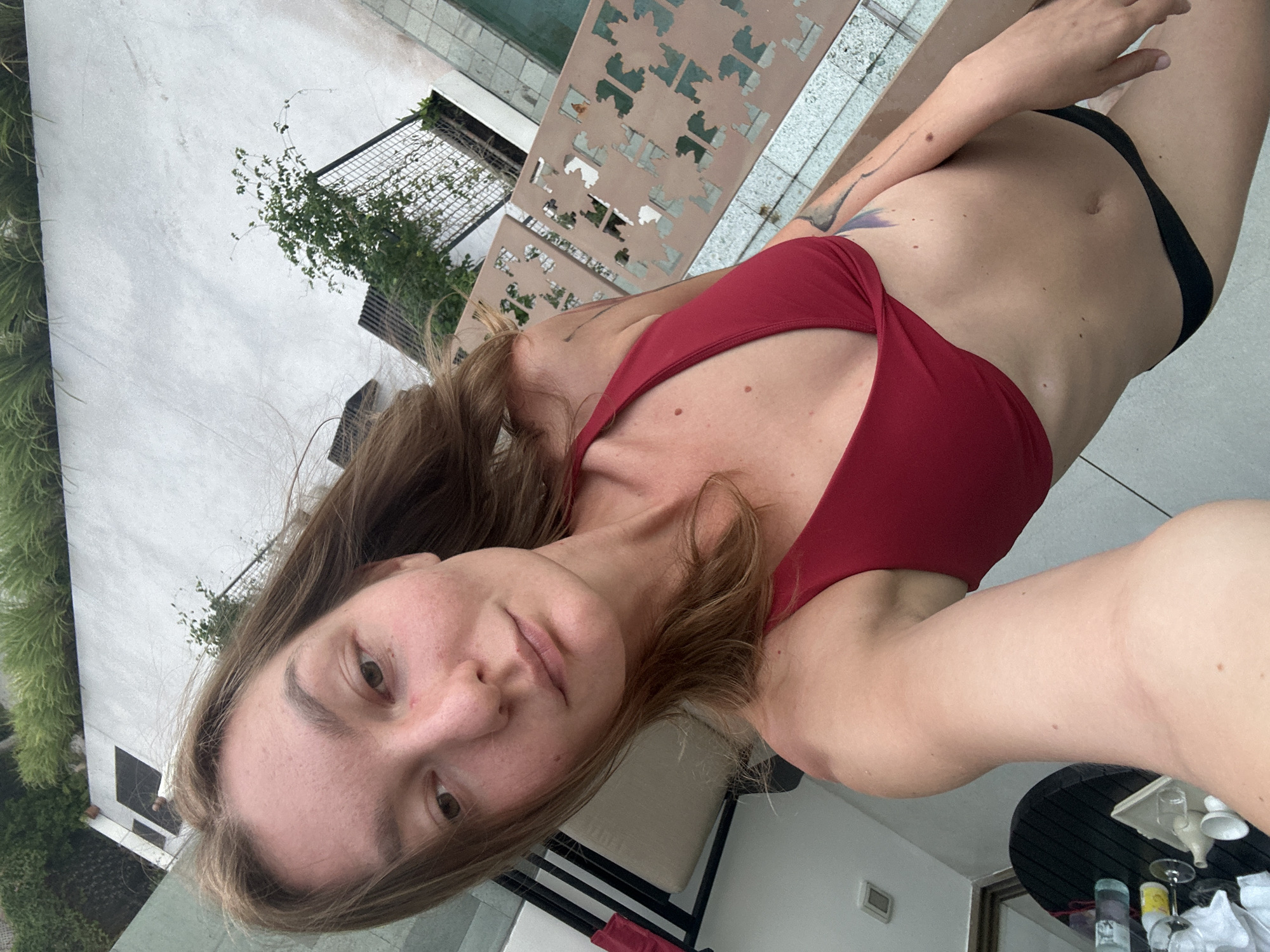 An outdoor selfie is taken by a fair-skinned person with long, light brown hair. The person is wearing a red bikini top and black bikini bottoms. They are standing on a tiled patio with a small pool visible in the background. To the left of the frame, there is a round table with drinks and a tray. Lush green foliage is visible in the upper right portion of the image. The lighting suggests it is daytime. The location is identified as Ko Samui District, Thailand.
