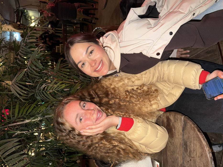 The image displays two young adult females seated at a round, dark wooden table within a dimly lit social establishment in Tel-Aviv, Israel, during evening or nighttime hours.
The female on the left has long, curly, light brown hair, a fair complexion, and visible freckles. She wears a tan puffer jacket over a red long-sleeved top. Her right hand is positioned against her jawline, while her left hand holds a smartphone displaying a messaging application with an active keyboard. A small, white and red box rests on the table to her left.
The female on the right has dark hair and wears a dark brown long-sleeved top, partially covered by a light beige puffer vest or jacket draped over her shoulders. Both females are smiling directly at the camera.
The background features dense green foliage, illuminated by warm artificial string lights. Several blurred figures of other patrons are visible in the mid-ground, seated at tables with high stools, indicating a public gathering space. The overall environment suggests an urban outdoor or semi-outdoor bar or restaurant setting.