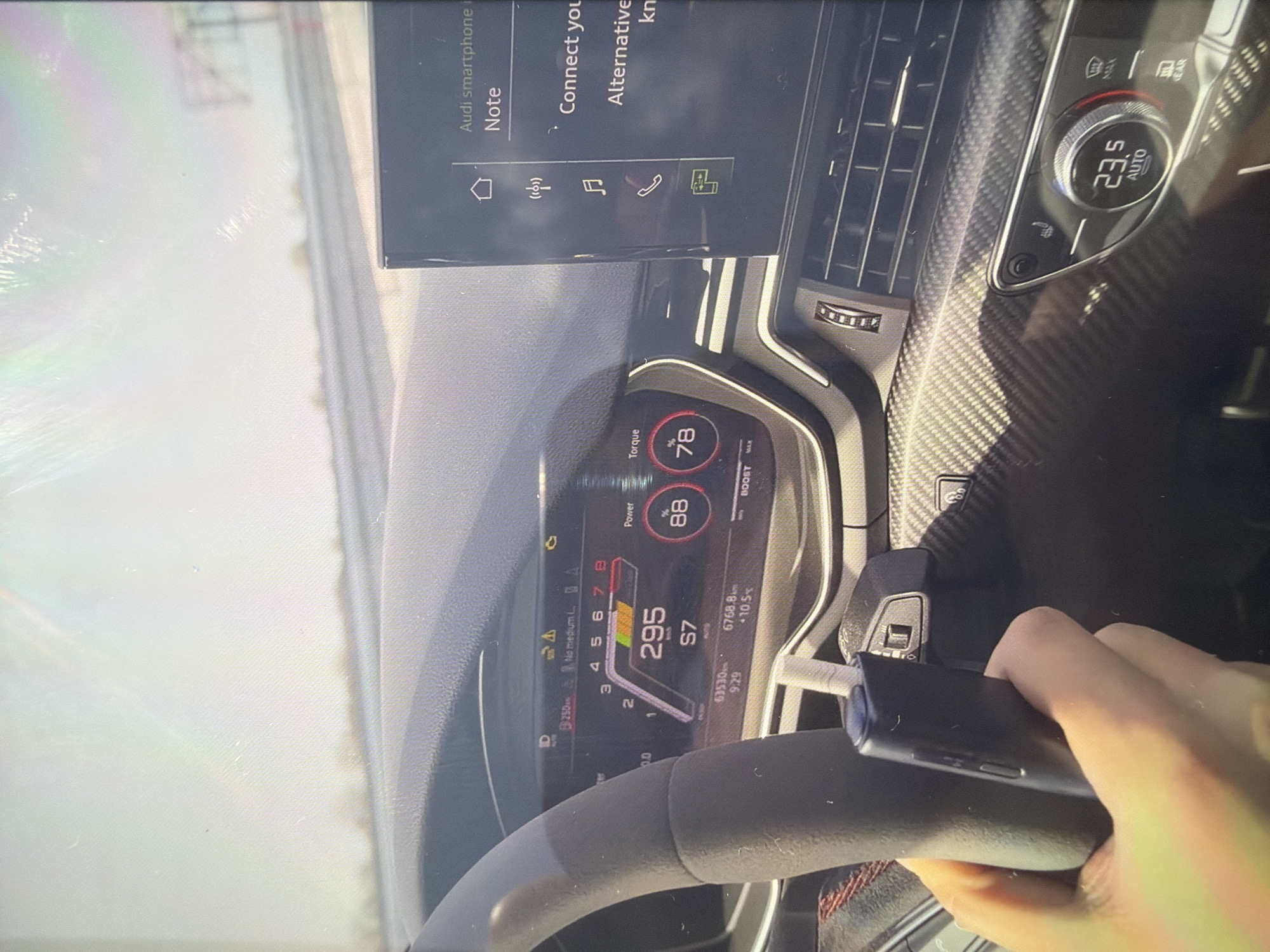The image captures a driver's perspective from inside a modern car, likely an Audi due to the "Audi smartphone interface" text on the central screen. A person's right hand holds a dark smartphone, resting on a steering wheel with red stitching.
The primary focus is on the car's digital instrument cluster, which displays a speedometer prominently showing "295 km/h," indicating a very high speed, and "S7," signifying the car is in 7th gear within a sport mode. Gauges for "Power" and "Torque" show values of 88% and 78% respectively, alongside a "BOOST" meter. The odometer reads "6768.8 km," and a trip meter shows "63530" and "9:29." A yellow warning triangle with an exclamation mark and text "No medium L.A." is also visible.
Above the instrument cluster, a central infotainment screen displays options related to the "Audi smartphone interface," including "Note," "Connect you," and "Alternative." Icons for home, wireless signal, music, phone call, and a vehicle function are present.
To the right, the climate control panel is visible, set to "23.5 AUTO," with a heated seat button and controls for "MAX" and "REAR" climate zones. The car's interior features carbon fiber trim on the dashboard, contributing to a sporty aesthetic. Bright daylight causes glare and reflections on the screens and windshield, suggesting it's daytime. The setting is identified as Dermantsi, Bulgaria.