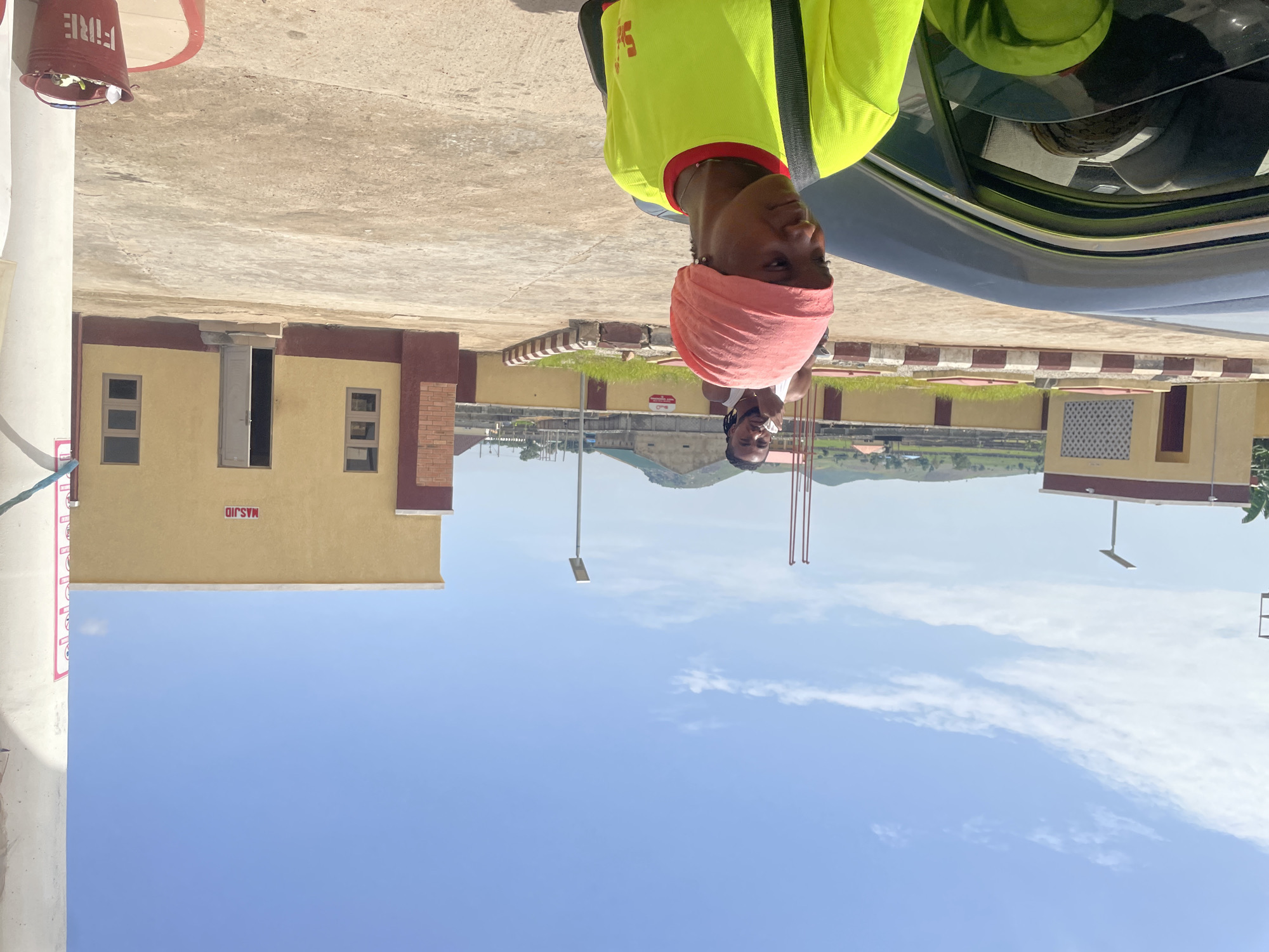 This is a vertical, rotated image taken in Pasere, Nigeria. The scene appears to be outdoors, possibly a street or open area adjacent to a building. The time of day is likely daytime, as indicated by the bright sky and clear lighting. The weather seems to be sunny with some scattered clouds.
In the foreground, a person is visible wearing a bright yellow shirt and a pink headscarf. They are looking towards the camera. Behind this person, another person can be seen, also looking towards the camera. They are positioned near a car.
The background features a building with yellow walls and dark red trim. This building has several windows and an open doorway. There is also a sign on the building with red text that appears to be in Arabic script. Further in the background, there are green fields and hills, suggesting a somewhat rural or semi-urban environment.
Other visible objects include a red bucket with the word "FIRE" written on it in white letters, attached to a white pole. There is also a lamppost visible.
The overall impression is a casual, everyday scene. The orientation of the image suggests it may have been taken with a phone held sideways.