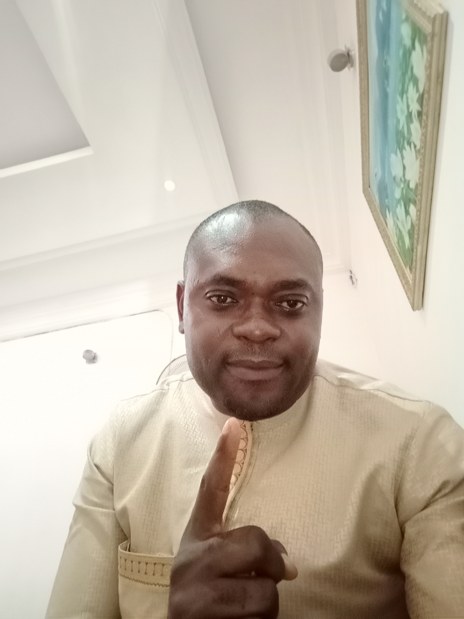 A dark-skinned man is positioned in a selfie-style photograph within an indoor setting. The man's head is shaved, and he is wearing a light tan colored, patterned tunic-style shirt with gold trim. His right hand is raised with his index finger extended upward. A painting in a gold frame hangs on the wall behind him to his right. A white door is visible behind him to his left. The ceiling is white with a recessed rectangular panel. The picture was taken in Gwarinpa, Nigeria.