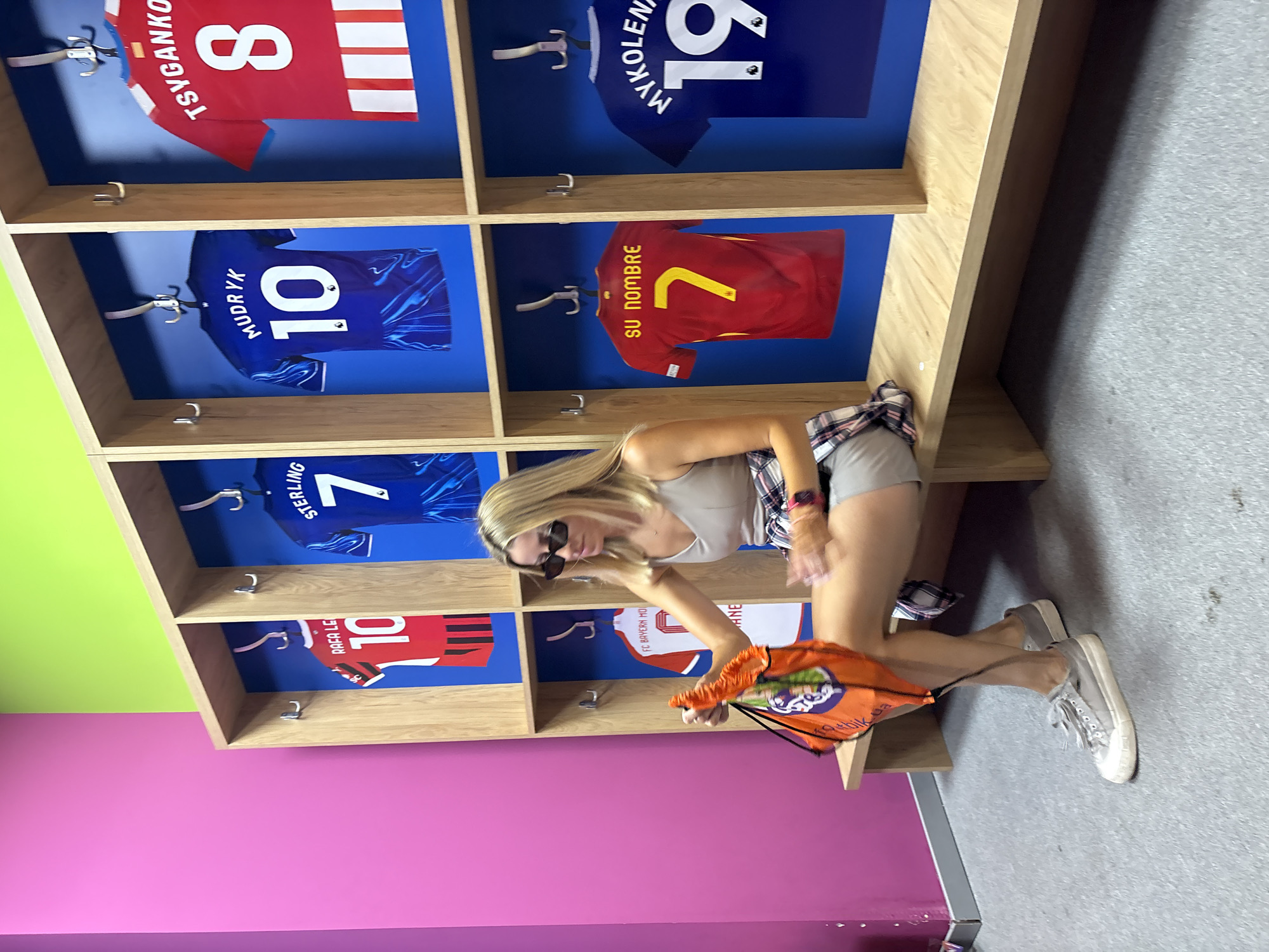 A blonde woman wearing a light-colored tank top, grey shorts, and grey sneakers is sitting on a wooden bench in a locker room. She is holding an orange drawstring bag with a white and green logo. She is wearing black sunglasses and a pink watch on her left wrist. The locker room features light wood cubbies with blue backdrops, each displaying a sports jersey. The jerseys are red, blue, and white, with names and numbers such as "RAFA LO 10", "STERLING 7", "MUDRYK 10", "TSYGANKO 8", "FC BAYERN MU", "SU NOMBRE 7", and "MYKOLEN 19". The walls are painted bright pink and lime green. The floor is grey carpet. The scene is consistent with a locker room at the Mestalla Stadium in Valencia, Spain.