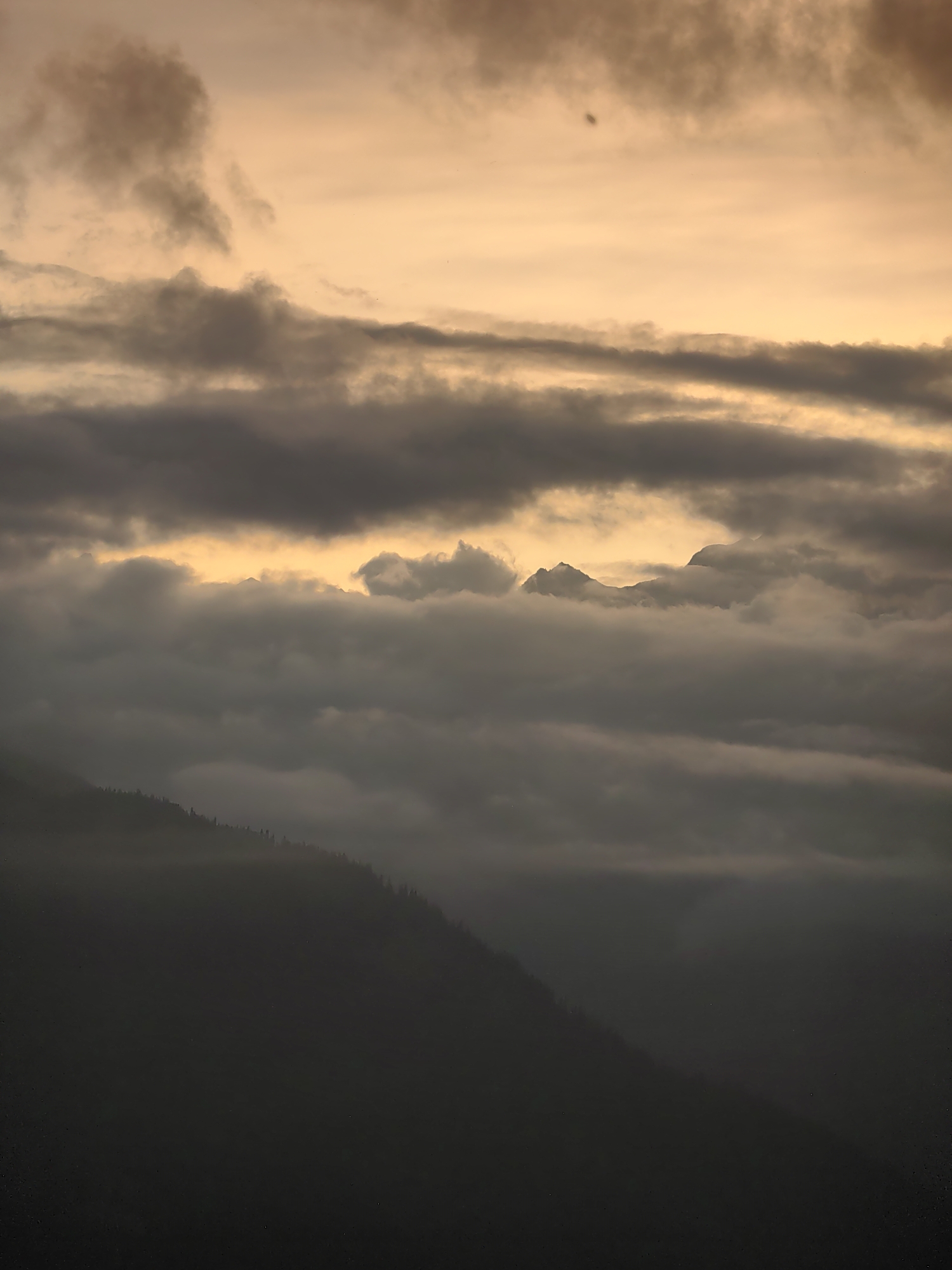 The image depicts a mountainous landscape with a dramatic sky. The foreground is dominated by a dark, silhouetted mountain slope covered in sparse trees, suggesting a steep incline. Above this, layers of thick, gray clouds obscure the view, with hints of lighter clouds and patches of golden light breaking through from behind. In the distance, the peaks of more mountains are visible, partially shrouded in mist and clouds, with a bright, hazy yellow light emanating from the horizon, indicating either sunrise or sunset. The overall atmosphere is moody and atmospheric, with a limited color palette of grays, blacks, and warm yellows. The specific location in the city of Aleo, India is not discernible from the visual elements present in the image.