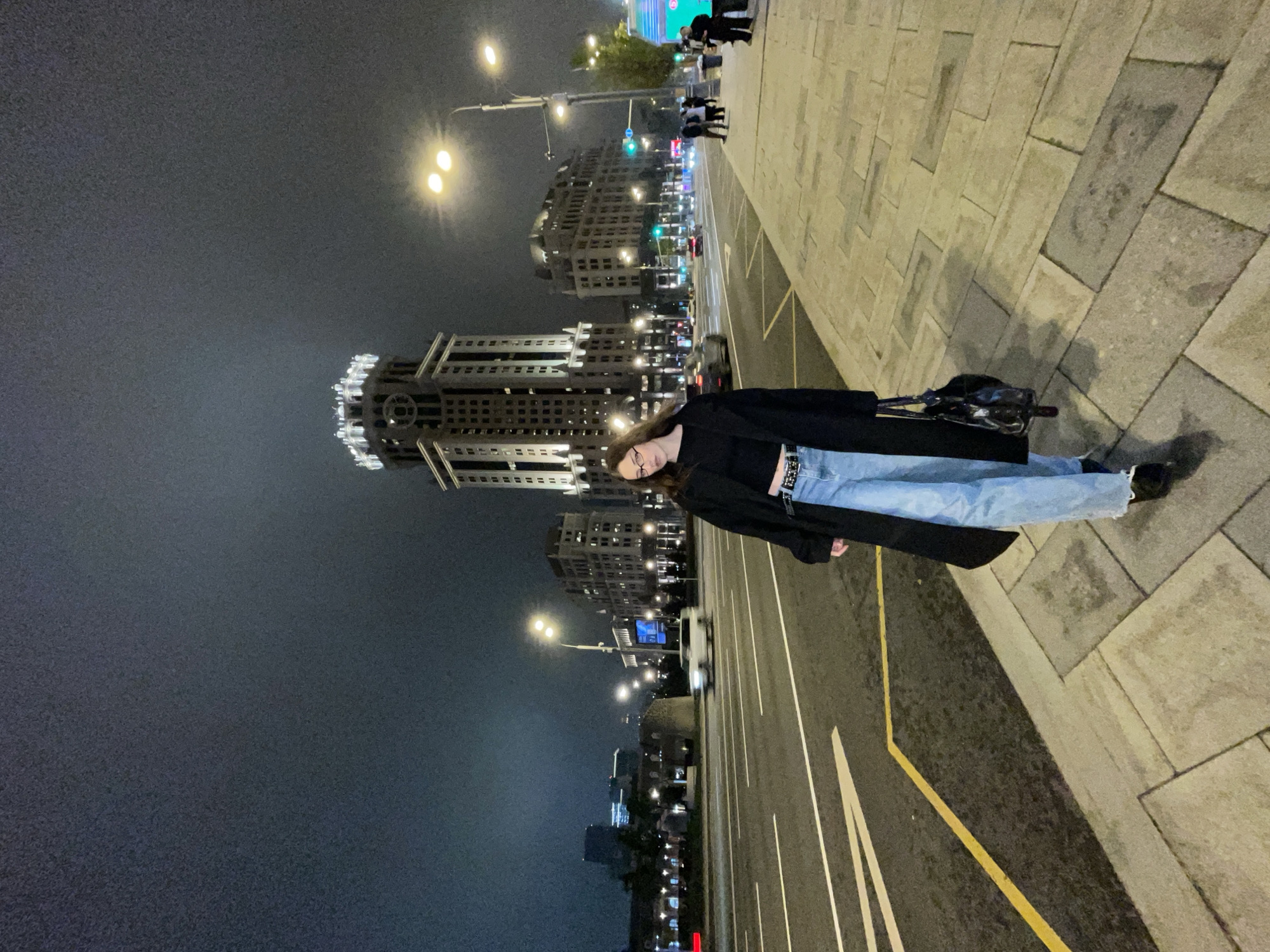 A vertically-oriented eye-level shot shows a person standing on the edge of a sidewalk at night with buildings in the background. The person is wearing a black coat, black shirt, light blue jeans, glasses, and black shoes. The person has a purse on their shoulder.
The person is standing on a sidewalk made of rectangular stones. Next to the sidewalk is a street with white and yellow lines on the ground. In the background are lit-up buildings of Moscow, Russia. Streetlights illuminate the buildings and the street. The sky is dark and cloudy.