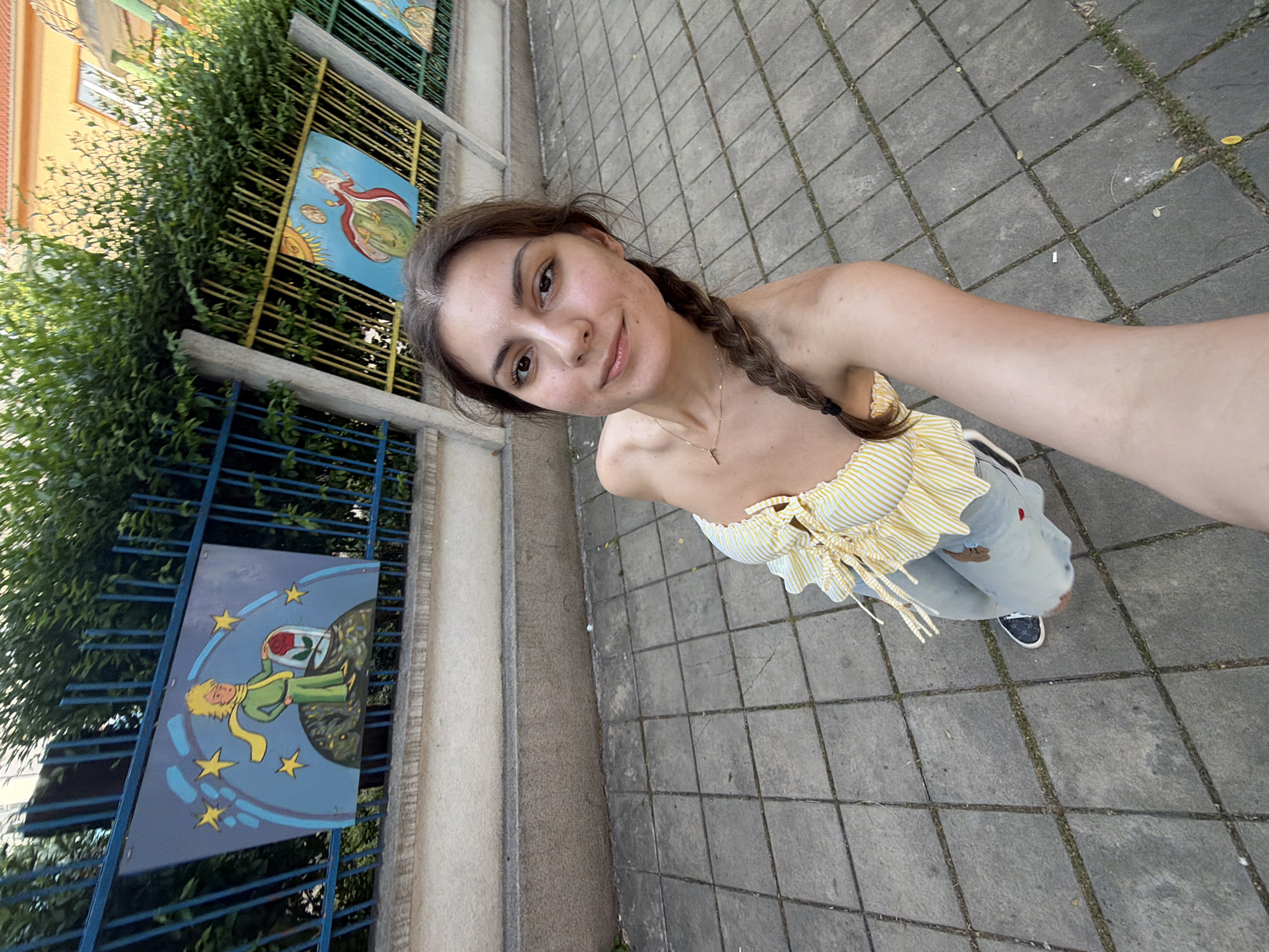 A young woman is captured in a sunlit outdoor selfie, likely taken in Burgas, Bulgaria. She stands at a slight angle, looking towards the camera with a gentle smile. Her brown hair is styled in a single braid, resting over her left shoulder. She wears a yellow and white striped, off-the-shoulder top, light-colored pants, and dark sneakers. A delicate necklace with a small pendant adorns her neck.
The setting features a paved ground with square concrete tiles. To her left, a fence is decorated with colorful, whimsical panels depicting characters and scenes from "The Little Prince" (Le Petit Prince). One panel prominently shows the Little Prince in his green suit, tending to his rose on a small planet amidst stars. Another panel portrays a figure, possibly an angel or another representation from the story, above a planet with a sun motif. Behind the fence, lush green foliage and a building with an orange wall are visible under what appears to be bright daytime sun, casting soft shadows. The overall atmosphere is pleasant and casual. No specific text is visible within the image itself.