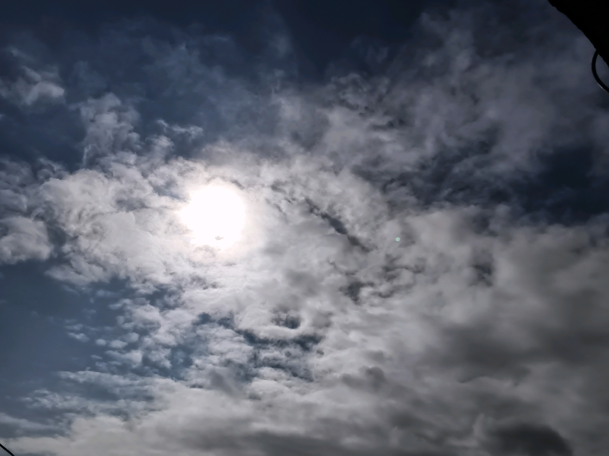 A sky photograph captures a dramatic interplay of sunlight and clouds. The sun, a bright, blown-out sphere, is partially obscured by wispy white clouds, creating a luminous halo effect. These bright clouds are juxtaposed against darker, more textured grey clouds that fill the majority of the frame, suggesting an approaching or receding storm. The overall lighting is harsh, with deep shadows cast by the denser cloud formations. No discernible landmarks of Thane, India, or any other specific geographical features are visible in this atmospheric composition. There are no people or discernible objects within the frame, only the dynamic patterns of the sky.