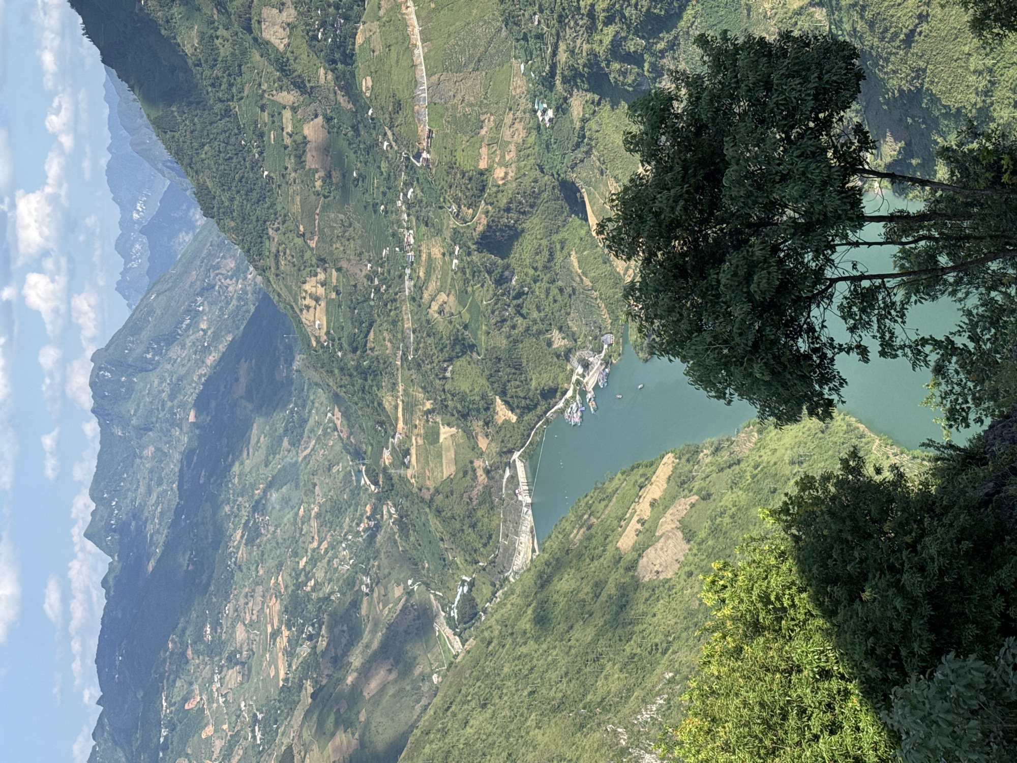 The image depicts a high-angle, scenic view of the landscape in Đồng Văn Commune, Vietnam. The rugged terrain is dominated by steep, green, mountainous slopes intersected by roads and villages. A river or reservoir is visible, with numerous boats docked near a structure, likely a dam or hydroelectric facility. The foreground includes foliage from trees, slightly obscuring the full panoramic view. Distant, hazy mountain ranges are visible in the background.