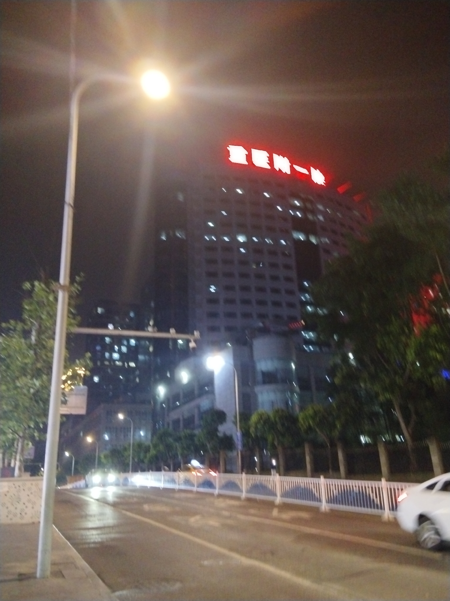 A nighttime street scene in Jiulongpo District, China, features a tall, illuminated building with red lettering on its facade. The building is surrounded by trees and other structures, some of which are also lit. A street lamp with a bright light source is prominently positioned in the foreground on the left. A white car is visible on the right side of the road, with its rear lights illuminated. The road appears wet, suggesting recent rain. A white fence with blue detailing separates the road from a sidewalk or pedestrian area. The sky is dark and hazy, characteristic of an urban night environment.
