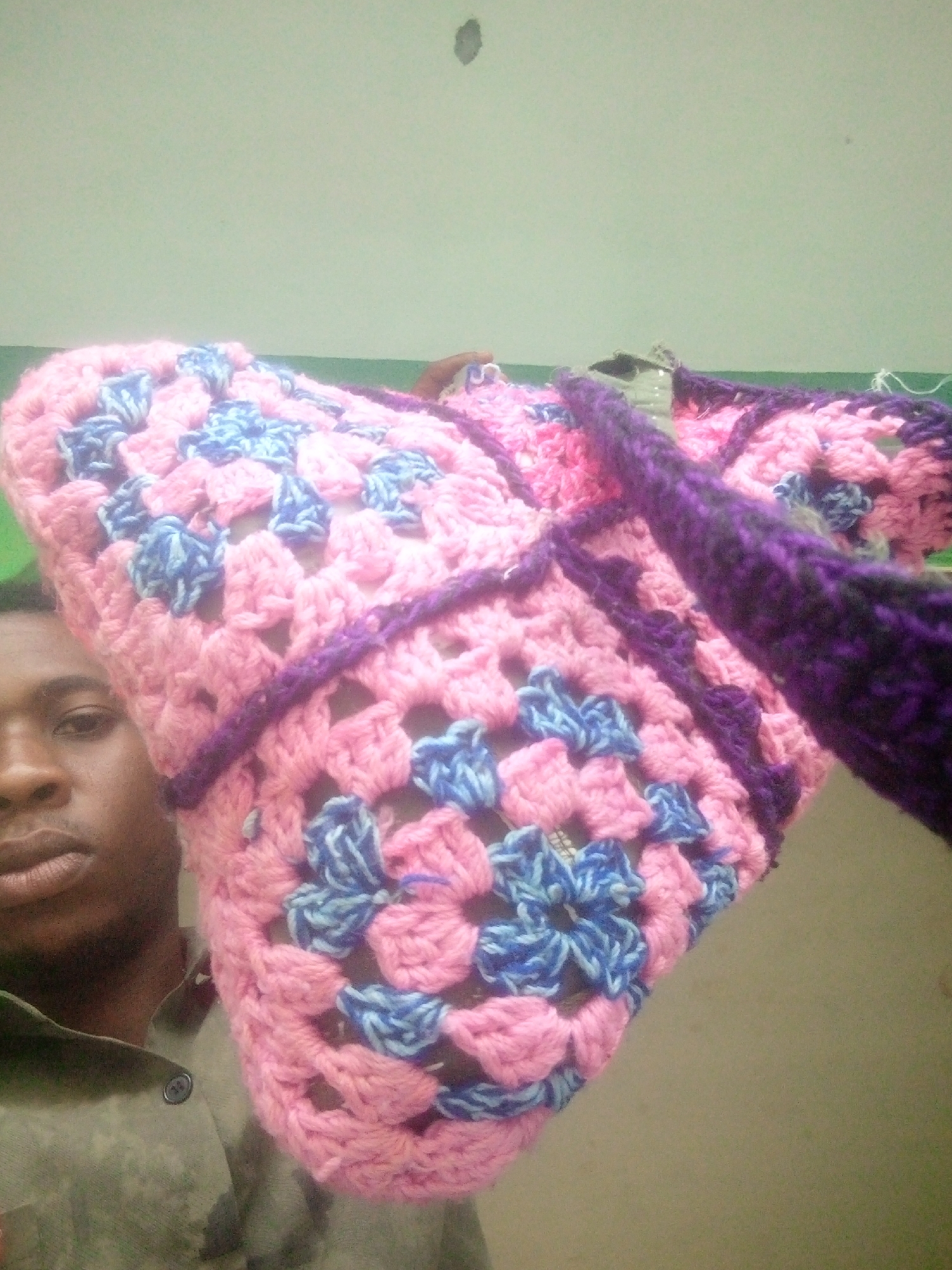 A man is holding up a pink and blue crochet bag with purple trim. The bag is in front of him, partially obscuring his face. The background is a plain, light green wall with a dark spot on it. The location is Unknown, Unknown.