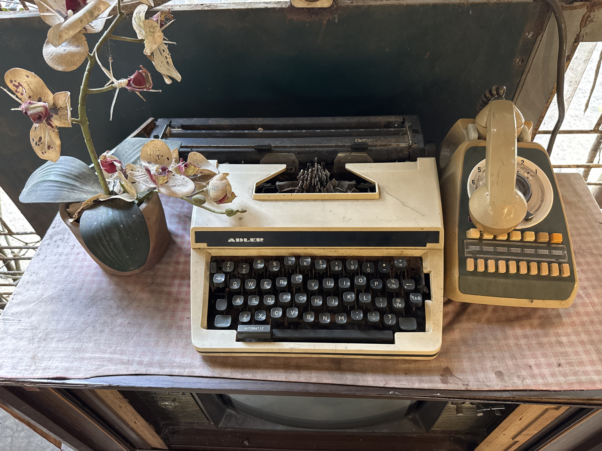 An overhead, medium shot captures an old Adler typewriter and a rotary dial telephone on a table covered with a pink checkered cloth. The typewriter is off-white with a black keyboard. The keys are visible, and the word "AUTOMATIC" is printed on a bar below the spacebar. The telephone is a light beige color with a dark green base. It has a coiled cord and a rotary dial. To the left of the typewriter, a vase holds a dried orchid arrangement with green leaves and brown and pink flowers. The background consists of a dark, metallic surface and a partial view of a wooden structure. The scene is likely set in an outdoor or semi-outdoor location in Cotbadatunong, Indonesia, as indicated by the presence of a window frame with a wire grate on the right side of the image. The overall impression is one of a vintage or nostalgic setting.