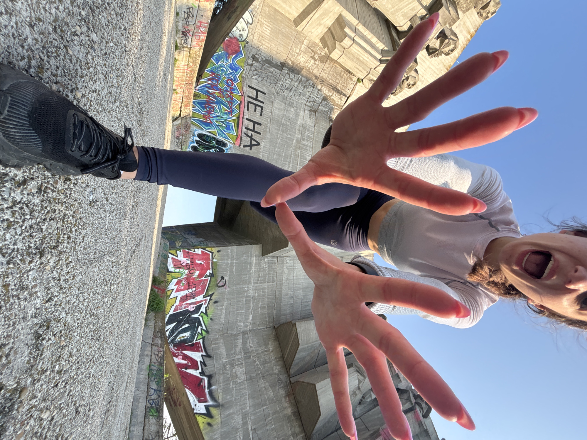 An extreme low-angle shot captures a person, likely female, posing playfully under a large concrete structure in Varna, Bulgaria. The individual is positioned almost horizontally across the frame, looking down at the camera with a wide-open mouth, suggesting a shout or laugh, and wide eyes. Both hands are extended towards the lens, fingers spread, creating a dramatic, wide-angle effect; her fingernails are painted pink. She is dressed in a light-colored, possibly white or light grey, long-sleeved athletic top, dark blue leggings, and black athletic shoes.
The setting appears to be an urban or industrial environment, possibly under a bridge or large overpass, characterized by massive, textured concrete structures. These structures are extensively covered in vibrant graffiti, featuring stylized letters in red, blue, green, and black. One distinct piece of graffiti reads "HE HA" in black block letters. The ground visible under her foot is a rough, pebbled surface. The clear blue sky in the upper right corner indicates daytime with bright sunshine. The overall mood is energetic and spontaneous.
