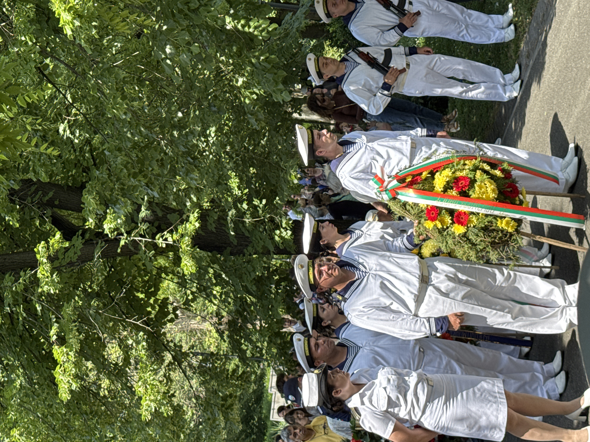 This media file depicts a formal outdoor ceremony, likely a memorial or commemorative event, held under bright daylight. A line of individuals in clean white naval uniforms, including both men and women, stands in an attentive posture, suggesting an honor guard or ceremonial participation. Many wear white peaked caps with insignia and have distinctive dark blue sailor collars. Some of the uniformed personnel further back are seen holding rifles.
Two large floral wreaths are prominent in the foreground. The most visible wreath features vibrant red and yellow flowers, including gerberas and chrysanthemums, and is adorned with ribbons in the colors of the Bulgarian flag: white, green, and red. Text on the white ribbon of this wreath is partially visible and appears to read "ОТ ОБЛАСТНА АДМИНИСТРАЦИЯ БУРГАС" (From Regional Administration Burgas), indicating the event's location and sponsor.
The setting includes lush green trees providing shade on the left side of the frame and a paved ground surface on the right. A crowd of civilian onlookers is visible behind the uniformed participants, observing the proceedings. The overall atmosphere conveyed is one of solemnity and respect, characteristic of such official ceremonies in Burgas, Bulgaria.