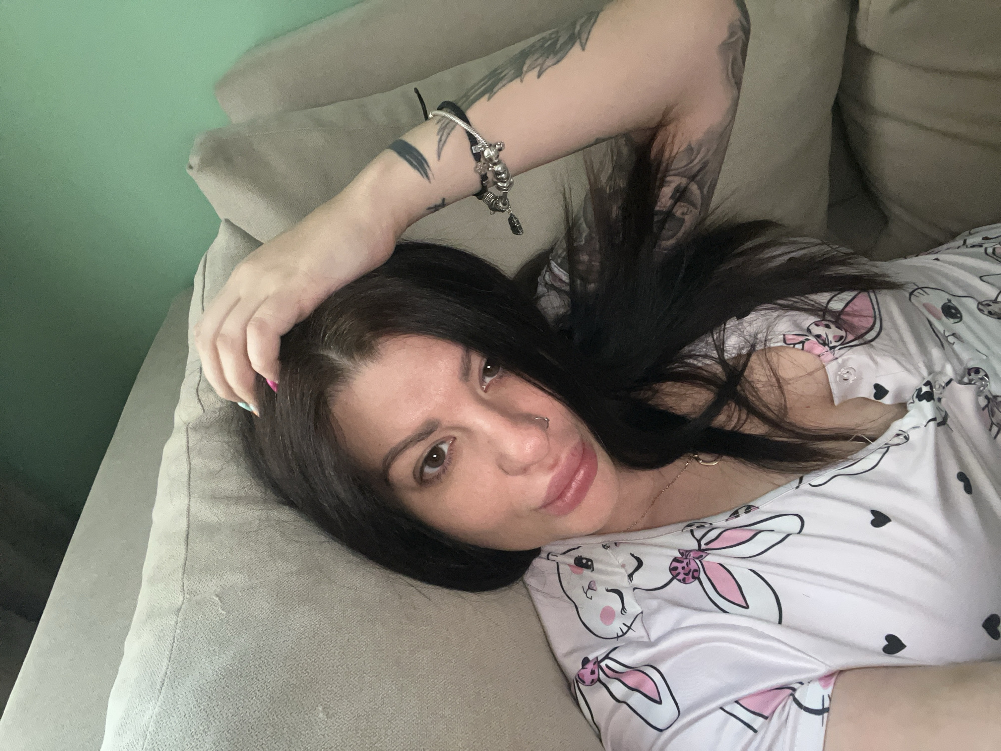 A woman with dark hair and tattoos on her arm is lying on a light-colored sofa. She is wearing a light-colored pajama top with a pattern of bunnies and hearts. Her hand is resting on her head. The wall behind the sofa is painted green.