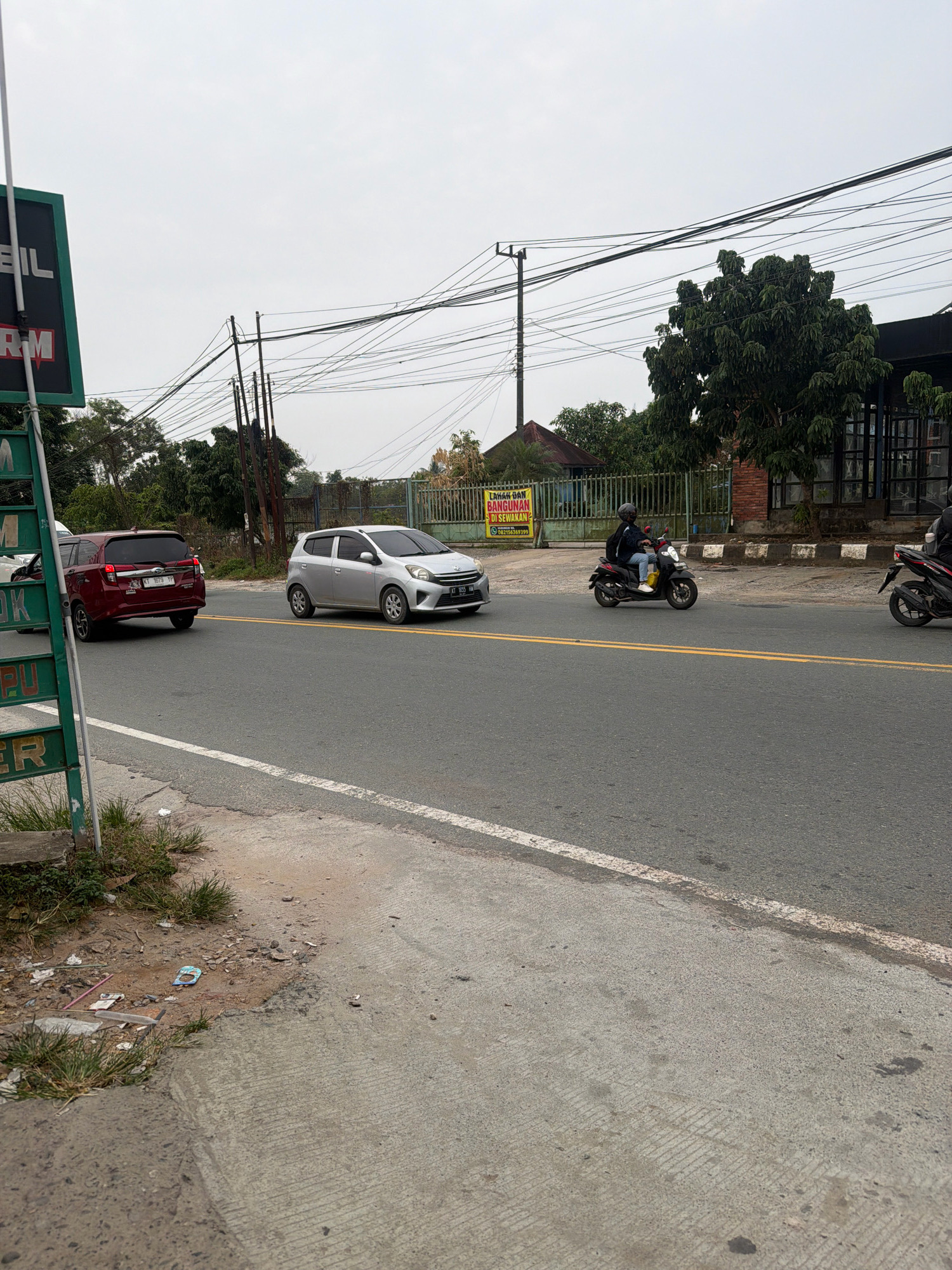 This image depicts a street scene in Balikpapan, Indonesia, likely on a cloudy day given the overcast sky. A paved road with a double yellow line marking the center runs horizontally across the frame. On the right side of the road, a dark-colored building with large windows and metal framing is visible. To the left of the building, a mature tree with dense foliage stands prominently. A fence runs along the edge of the property.
In the foreground and on the road, several vehicles are present. A red SUV is traveling away from the camera on the left lane. A silver hatchback is also moving on the road, further ahead and in the right lane. Two motorcycles are on the road as well; one is closer to the camera and the other is further ahead. The motorcycle closer to the camera has two occupants, a driver and a passenger, both wearing helmets. The environment appears to be urban or suburban, with power lines and poles visible overhead. A large green sign with white lettering is positioned on the far left of the image, partially obscuring the view of the vehicles and roadside. The roadside on the left is unpaved and shows some scattered debris and vegetation. The overall impression is a typical street view in a developing area.