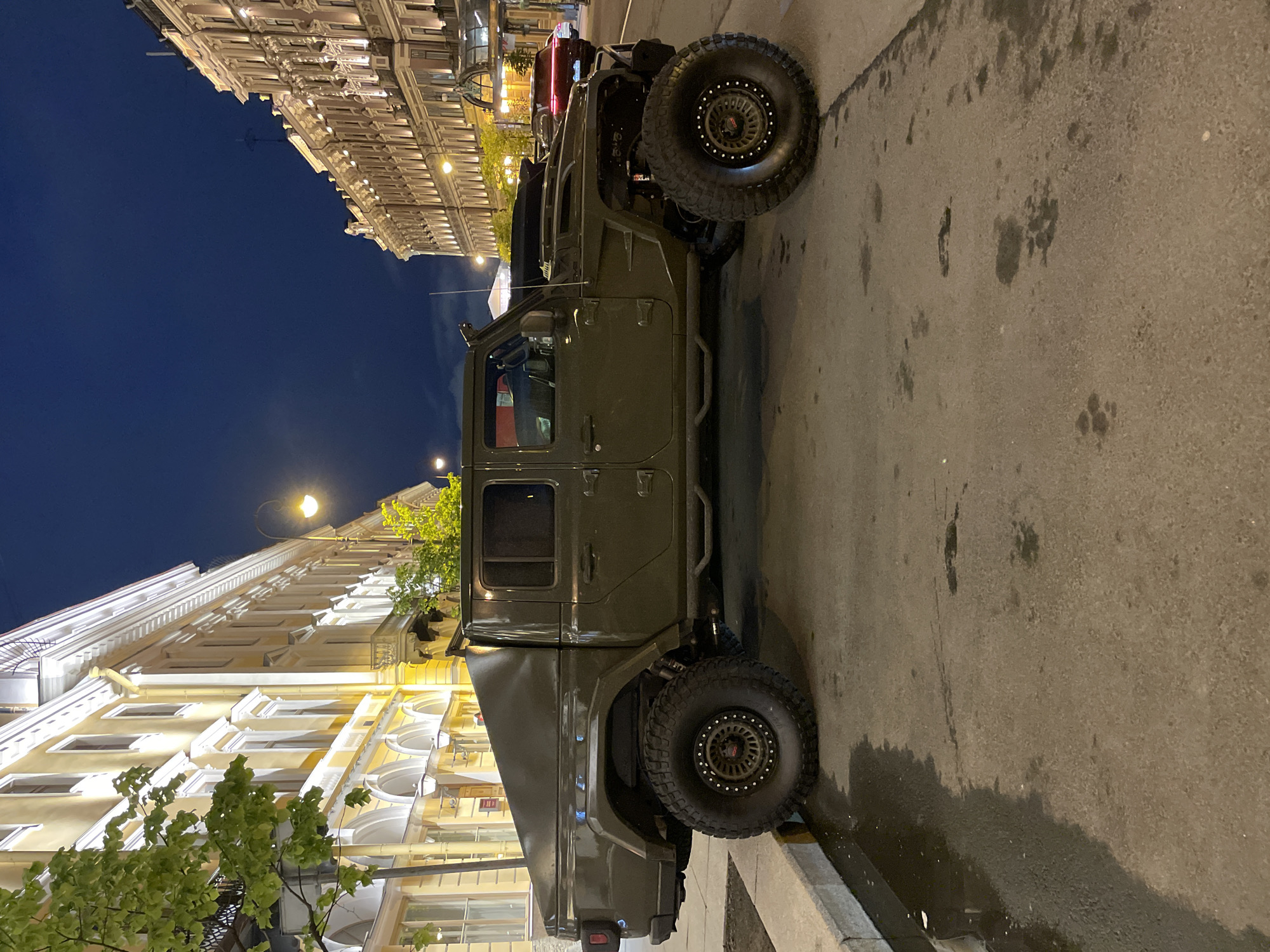The image shows a dark green military-style vehicle parked on a street in Saint Petersburg, Russia. The vehicle has large, off-road tires and appears heavily modified. Buildings line the street, illuminated by streetlights, and it is night. The sky is dark, and the scene suggests an urban environment at dusk or night. There are trees with green leaves along the left side of the image. The street surface is wet and shows signs of wear.