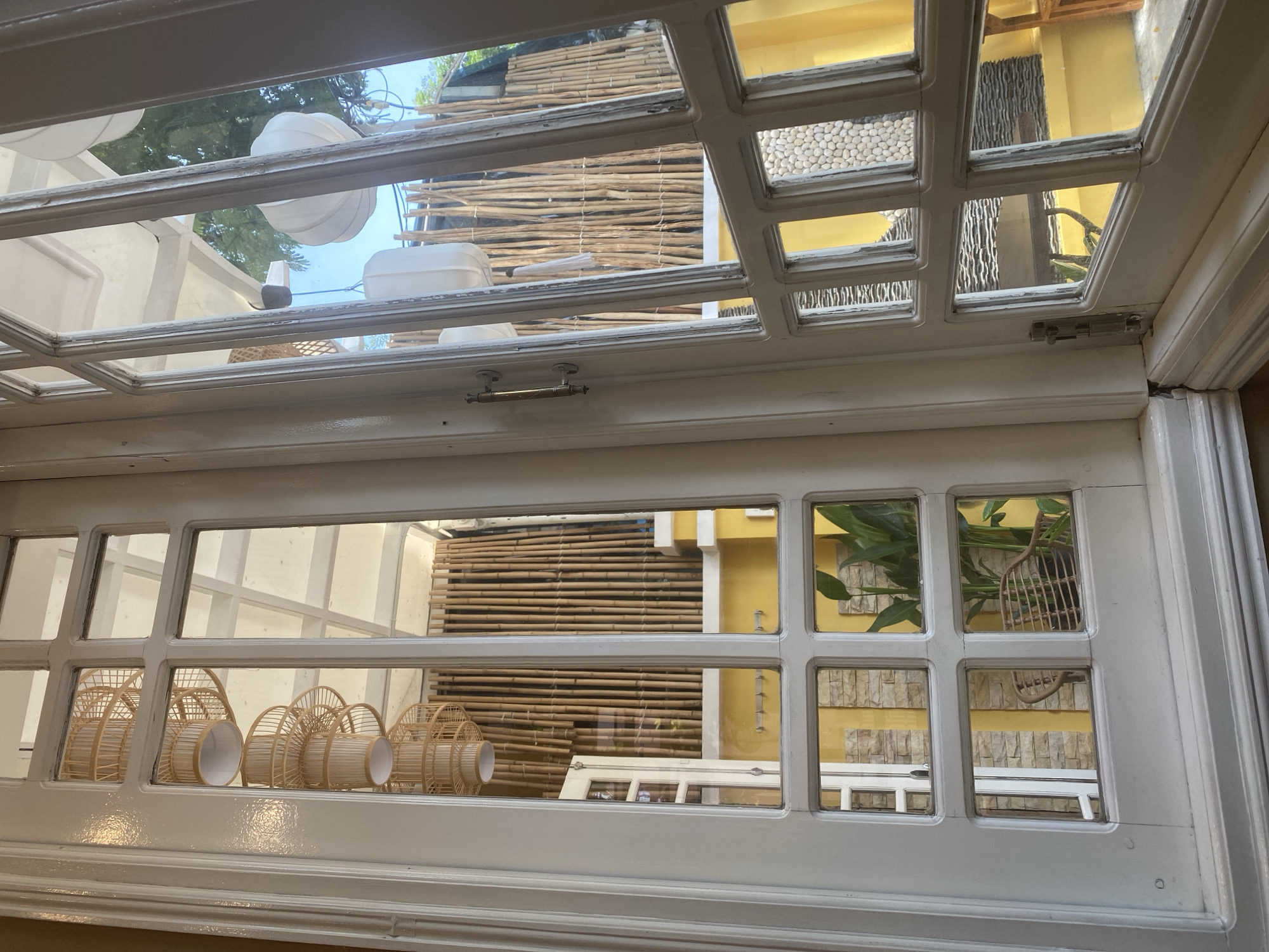 The image shows a view through a white-framed glass door, likely in Da Nang, Vietnam, given the architectural style and outdoor elements. The door is divided into multiple panes, some of which reflect the interior, while others offer a view of the exterior.
The reflection in the upper left panes shows a stack of decorative pendant lights, possibly made of woven bamboo or straw, with a white cylinder inside each. These lights are hanging from the ceiling.
The exterior view, seen through the right-hand panes, reveals a rustic outdoor area. A wall constructed of vertical bamboo poles serves as a backdrop. White, lantern-like light fixtures are visible hanging from an overhead structure. Trees and bright blue sky are also present in the background, suggesting an outdoor setting such as a patio or garden.
In the lower panes, reflections show elements of the interior again, including a yellow wall, stone tiling, and a wicker chair. One pane shows a large green plant.
The overall impression is one of a bright, airy space with a tropical or natural aesthetic, possibly a cafe, restaurant, or a residence with a courtyard. The light appears to be natural daylight. No people are visible in the image. The door itself has a metallic handle and a bolt on the right side.