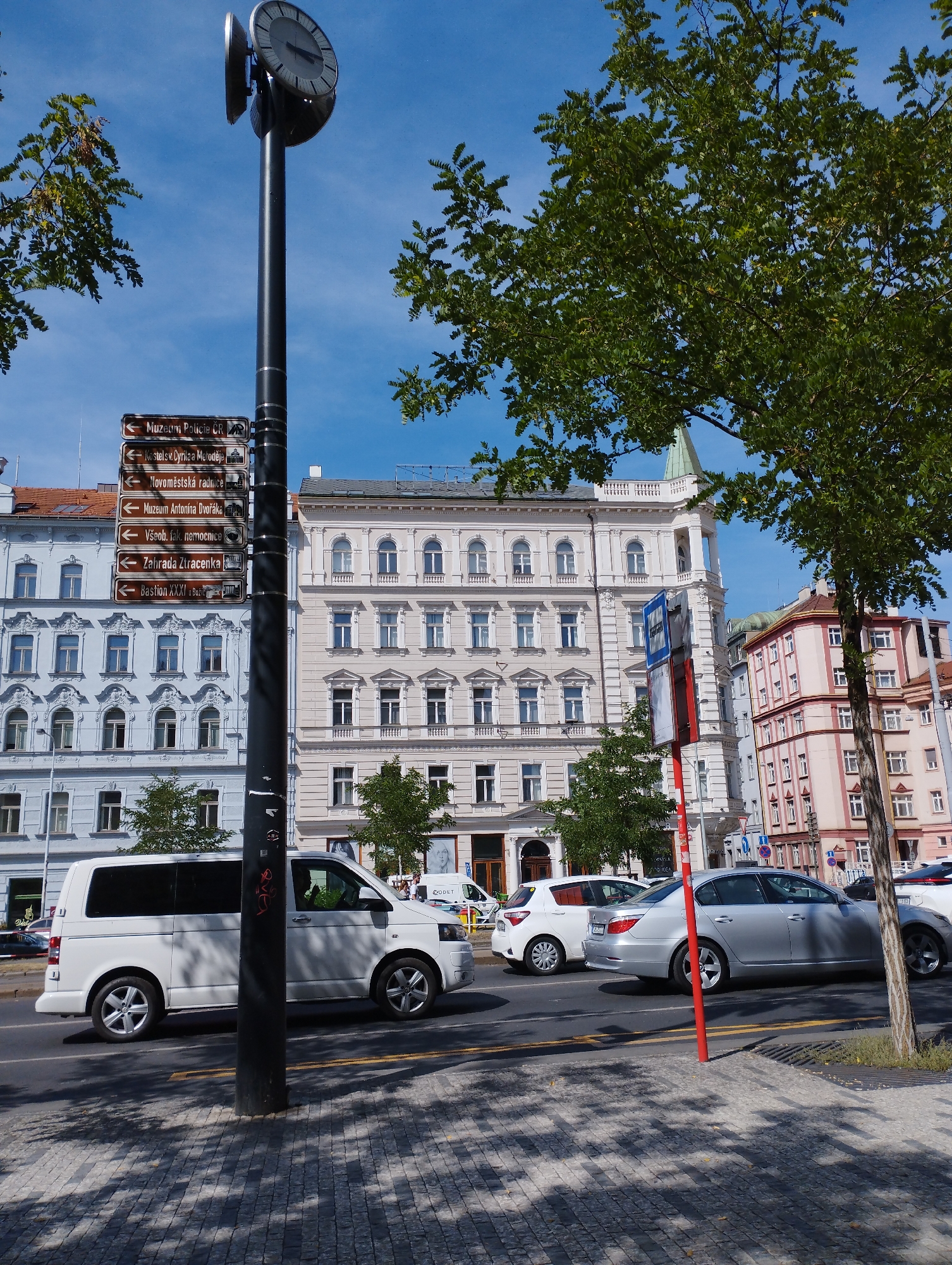 The image shows a street scene in Prague, Czechia. In the foreground, a large black lamppost with a clock face is positioned on a cobblestone sidewalk, with trees casting shadows across the pavement. Several directional signs with white text on brown backgrounds are attached to the lamppost, indicating directions to various landmarks such as "Muzeum Policie ČR" and "Národní technické muzeum." To the right of the lamppost, a red and blue bus stop sign is visible.
In the street, several vehicles are present. A white van is driving in the left lane, while a silver sedan is in the right lane. Further down the street, other cars and buildings are visible. The background features a row of ornate, multi-story buildings with classical architectural details, painted in light blue, cream, and pink. The sky is clear and blue. The overall scene depicts a busy urban environment with historical architecture and contemporary traffic.