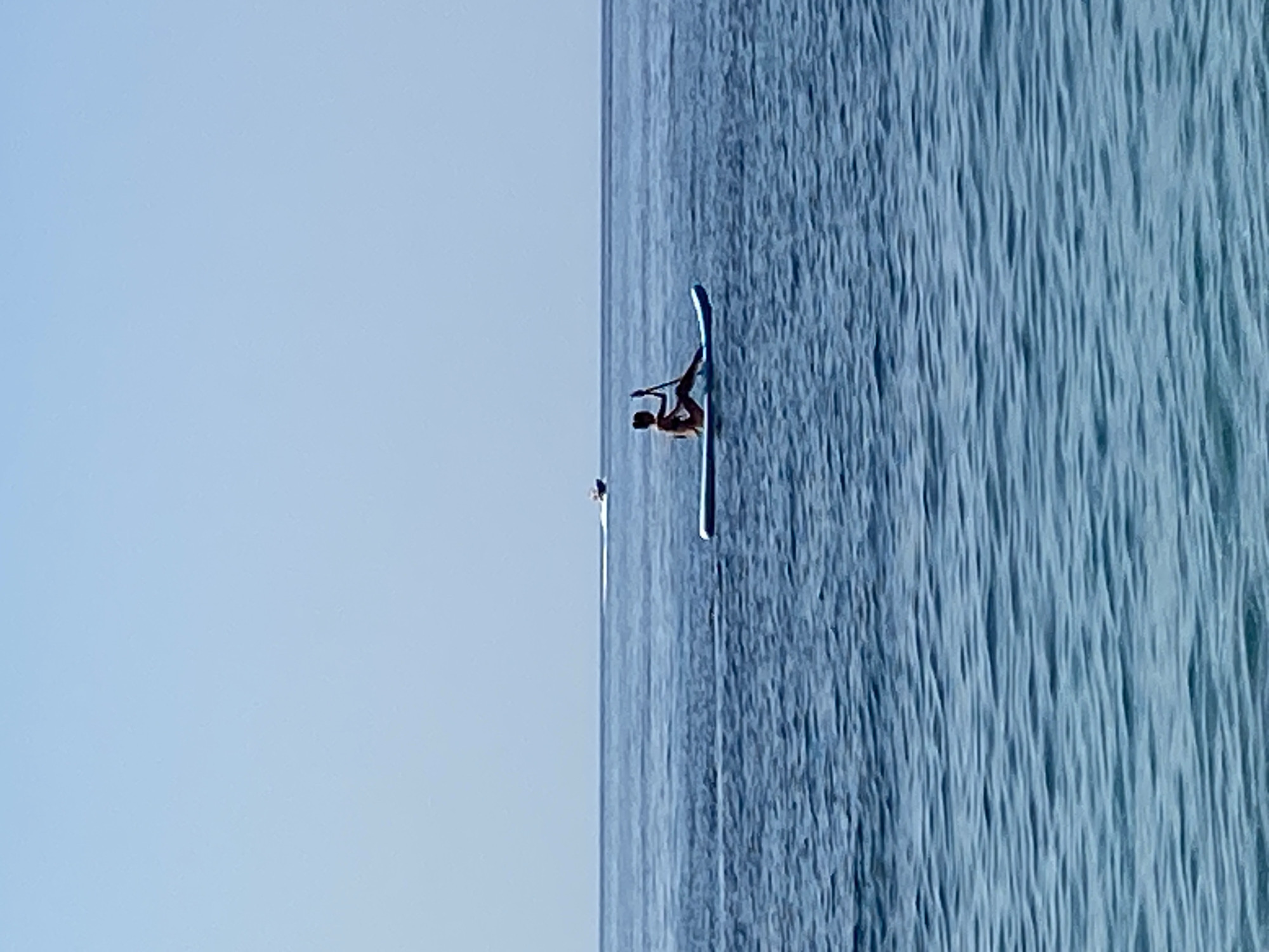 A lone person is paddleboarding on the Adriatic Sea, with the coast of Prijevor, Montenegro, visible in the distance. The person is sitting on a blue paddleboard, holding a paddle. The sea is calm and a boat is visible in the far distance. The sky is clear and blue.