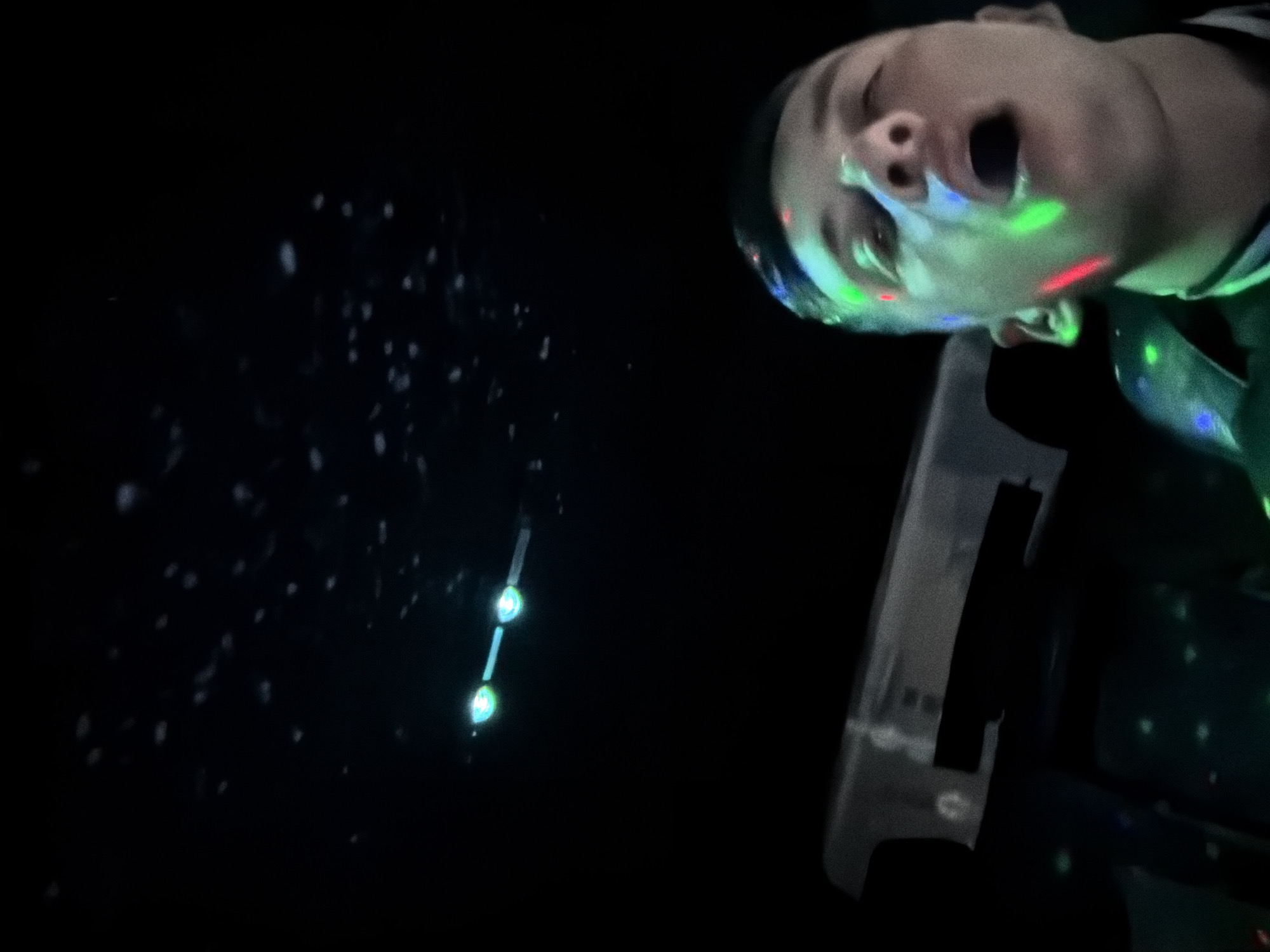 A tilted, low-light photograph captures a person's face illuminated by vibrant disco lights in a dark environment. The individual, appearing to be a child or young person, is positioned in the upper right quadrant of the image, looking upwards or sideways with their mouth wide open, suggesting singing, shouting, or an expression of excitement. Red, green, and blue light patterns are projected onto their face, highlighting features and casting colorful reflections.
The background is predominantly black. In the mid-left, three distinct circular lights, emitting a greenish-blue glow, are arranged vertically. Numerous small, scattered white or light grey specks are visible across the dark space, resembling dust particles, water droplets, or light bokeh. A partial view of a light-colored, possibly structural or furniture-like object, is visible in the bottom right.
The overall setting appears to be an entertainment venue, such as a party, club, or laser tag arena, characterized by the dynamic colored lighting and darkened room. The time of day is clearly night or a similarly dark indoor situation. The person's expression and the festive lighting evoke a sense of engagement and enjoyment. There is no visible text in the image.