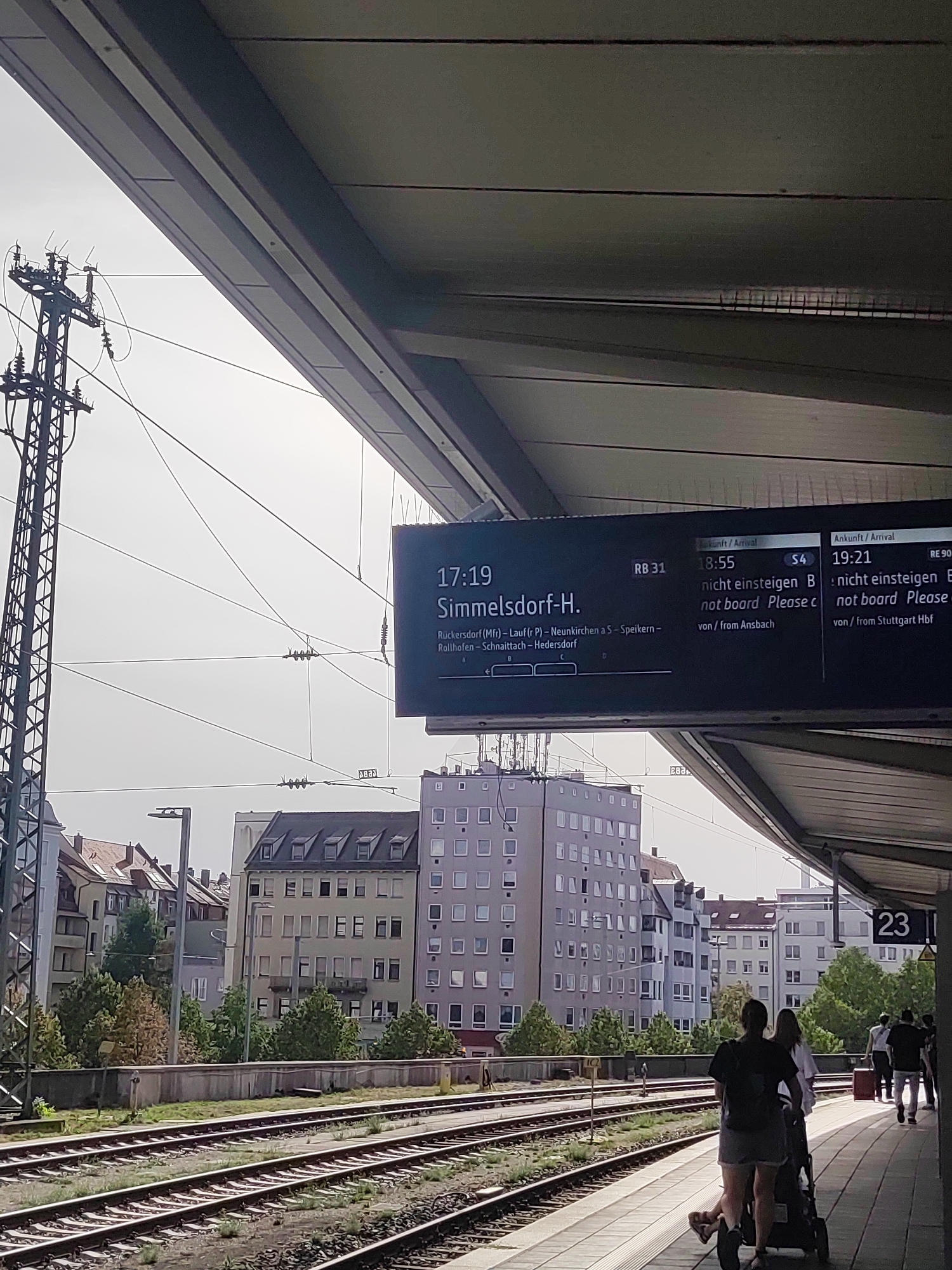The image captures a view from a covered train station platform in Nuremberg, Germany. In the foreground, train tracks run parallel to the platform. In the mid-ground, several multi-story buildings are visible, characterized by their windowed facades and architectural styles common to urban European settings. The sky is overcast. A digital display board mounted on the platform shows train departure information, including times, destinations, and platform numbers. The display indicates departures for Simmelsdorf-H., and other destinations from platforms RB 31 and RE 90. Several people are present on the platform, with one individual pushing a stroller and another couple walking away from the camera. The overall scene depicts a typical day at a public transportation hub.
