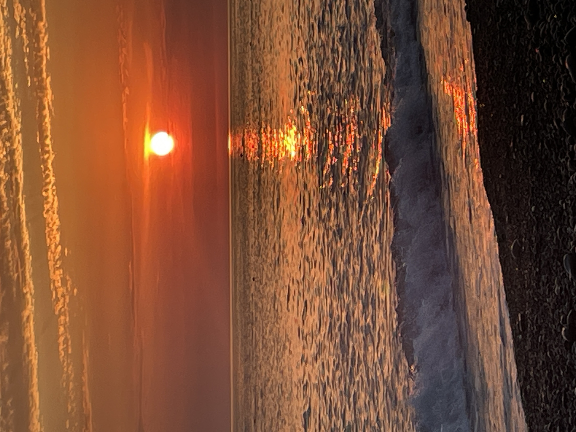 A sunset over the ocean is depicted. The sun, a bright white orb with a yellow halo, is positioned near the center of the frame, partially obscured by thin, elongated clouds streaked with orange and yellow. The ocean's surface is a shimmering expanse of orange and gold, reflecting the sun's light. Gentle waves are breaking on a dark, pebble-strewn beach in the foreground. The sky transitions from a deep orange near the horizon to a softer pinkish-orange at the top. The overall mood is serene and picturesque.