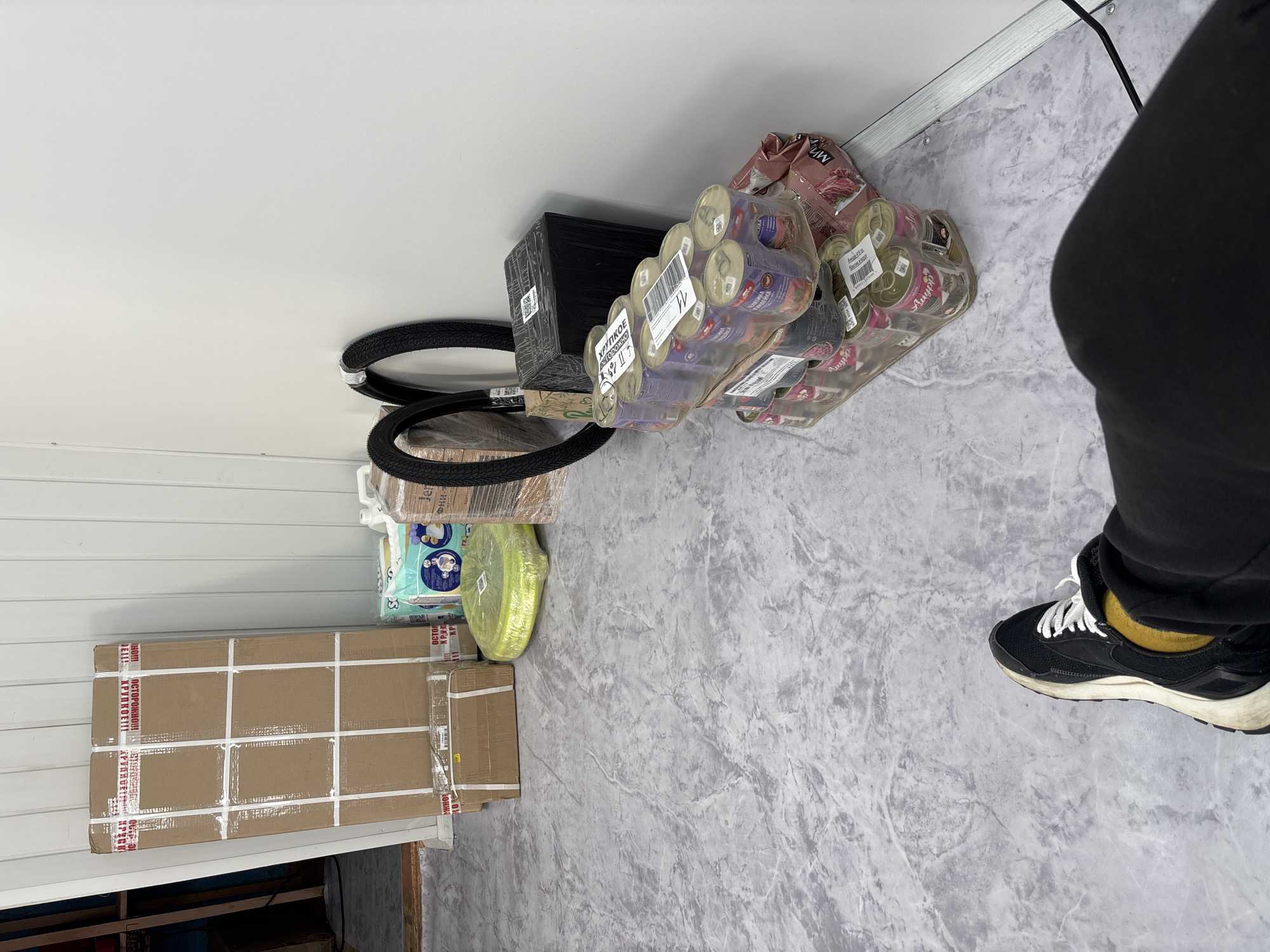 The image depicts a cluttered storage area, possibly a warehouse or a back room, with various items stacked and placed on a marble-patterned floor. In the foreground, a person's legs and feet are visible, wearing black pants and sneakers with white laces and a yellow sock. The person is seated, suggesting a moment of rest.
Dominating the background are several large cardboard boxes, some sealed with white tape and red warning labels. One particularly tall box is leaning against a white corrugated wall. To the right of the boxes are two bicycle tires, one partially wrapped in plastic.
In the mid-ground, a stack of canned goods, likely pet food based on the labels visible on some cans (e.g., "АППП"), is arranged in a plastic-wrapped pallet. Next to the cans is a black, rectangular object, possibly a speaker or storage unit, wrapped in plastic. A light green, coiled object, possibly a hose or cable, is also present. A package of diapers is visible between the large cardboard boxes and the tires.
The environment appears to be indoors, with a white corrugated wall forming the backdrop. The lighting is even, suggesting artificial illumination. The overall impression is one of storage or waiting for delivery/shipment. There is no direct indication of the city of Vileyka, Belarus within the image itself; this information would need to be provided externally to contextualize the scene.