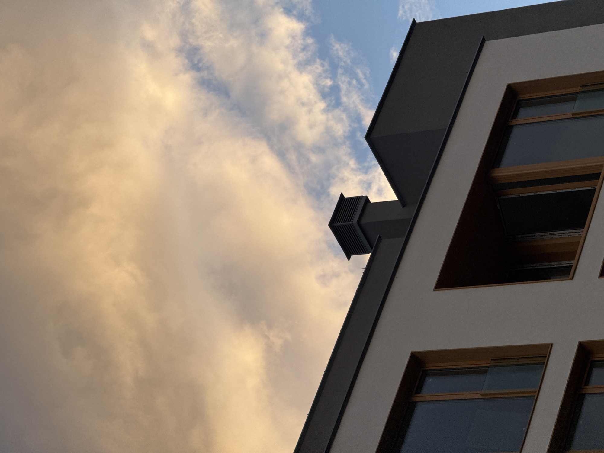 A low-angle shot captures the exterior of a modern building with geometric lines under a sky of orange-tinted clouds and patches of blue. The building's facade features large, recessed windows with wooden frames, and a dark grey trim accentuates its edges. An air vent or chimney projects from the side of the building. The image composition emphasizes the building's architecture against the dynamic sky. The photo was taken in the city of Sofia, Bulgaria.