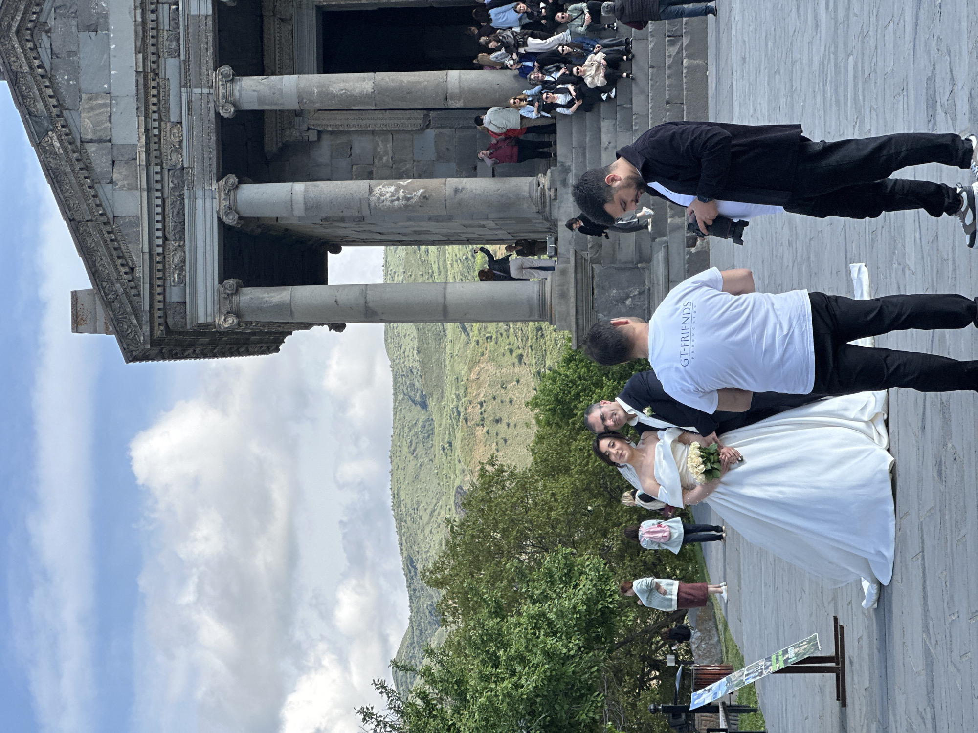 The image captures a wedding photoshoot at the Temple of Garni in Garni, Armenia. A bride in a white gown and a groom in a suit stand together on a stone terrace with a mountain visible in the background. Two photographers are present, one dressed in black with a camera and another in a white t-shirt with "GT-FRIENDS PRODUCTION" printed on it, appearing to direct the couple. Behind the couple, there are some people, green trees, and an information stand. The temple's stone columns and part of the upper structure are visible at the top of the frame. People are visible on the steps leading up to the temple. The sky above is blue with scattered clouds.