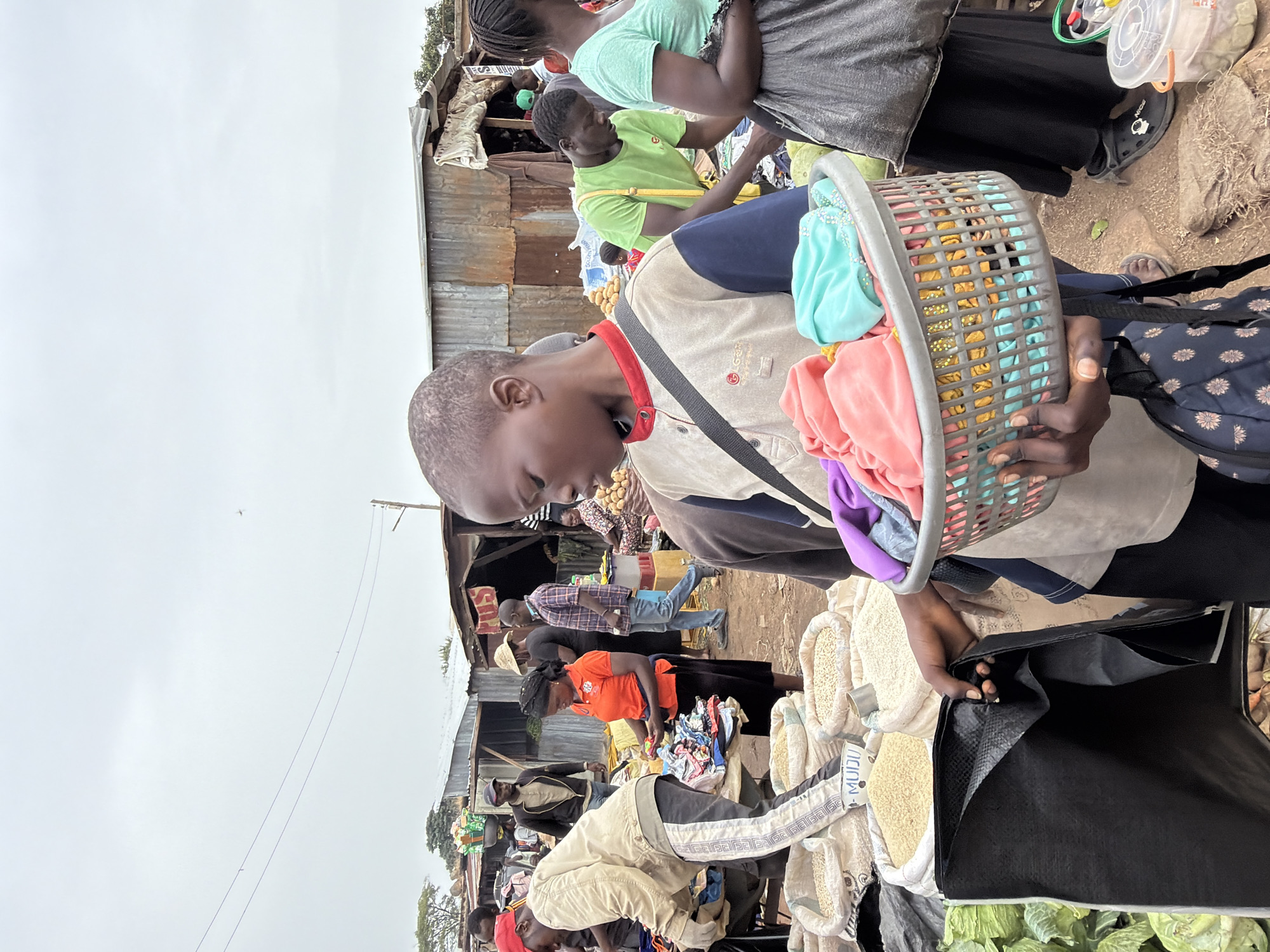 The image shows a busy market scene in Gura Top, Nigeria. A young man in a light-colored shirt with dark blue sleeves and a shoulder bag is holding a plastic basket filled with colorful clothes. He appears to be shopping, as he's looking at sacks of what appear to be grains or beans. Other people are visible in the background, some shopping, others standing around. There are stalls covered with corrugated metal sheets, possibly market stands, with various goods displayed. There are vegetables and other goods displayed on the ground. The sky overhead is overcast.