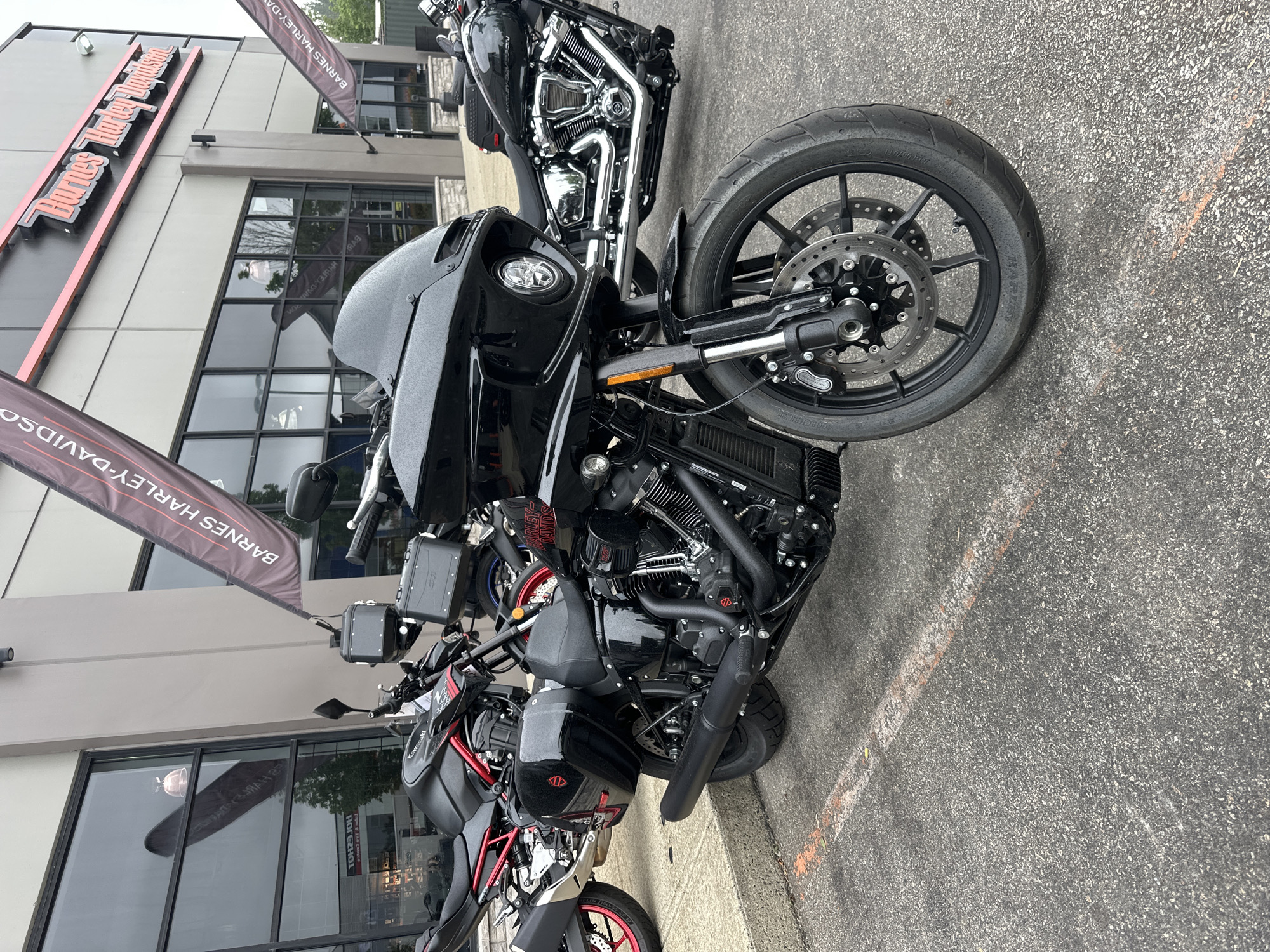 This media file captures a close-up, tilted view of several black motorcycles parked outside a Harley-Davidson dealership. The dealership's building is visible in the background, with large windows and a prominent sign reading "Barnes Harley-Davidson" in orange lettering. Two large banner flags with "BARNES HARLEY-DAVIDSON" printed on them are also present, one hanging from the building and another in the foreground.
The motorcycles are the primary subjects. The front of a large touring motorcycle is prominently featured, with its black fairing, headlight, and wheel. To its right, another motorcycle, a sportbike with red accents on its wheels, is partially visible. The motorcycles appear to be parked on an asphalt surface, with faint orange and white lines suggesting a parking area.
The scene appears to be outdoors during daylight hours, with overcast or bright sky reflected in the building's windows. The overall impression is one of a commercial setting, likely a dealership showcasing its inventory. There are no people visible in the image. The focus is on the machines themselves and the branding of the dealership.
