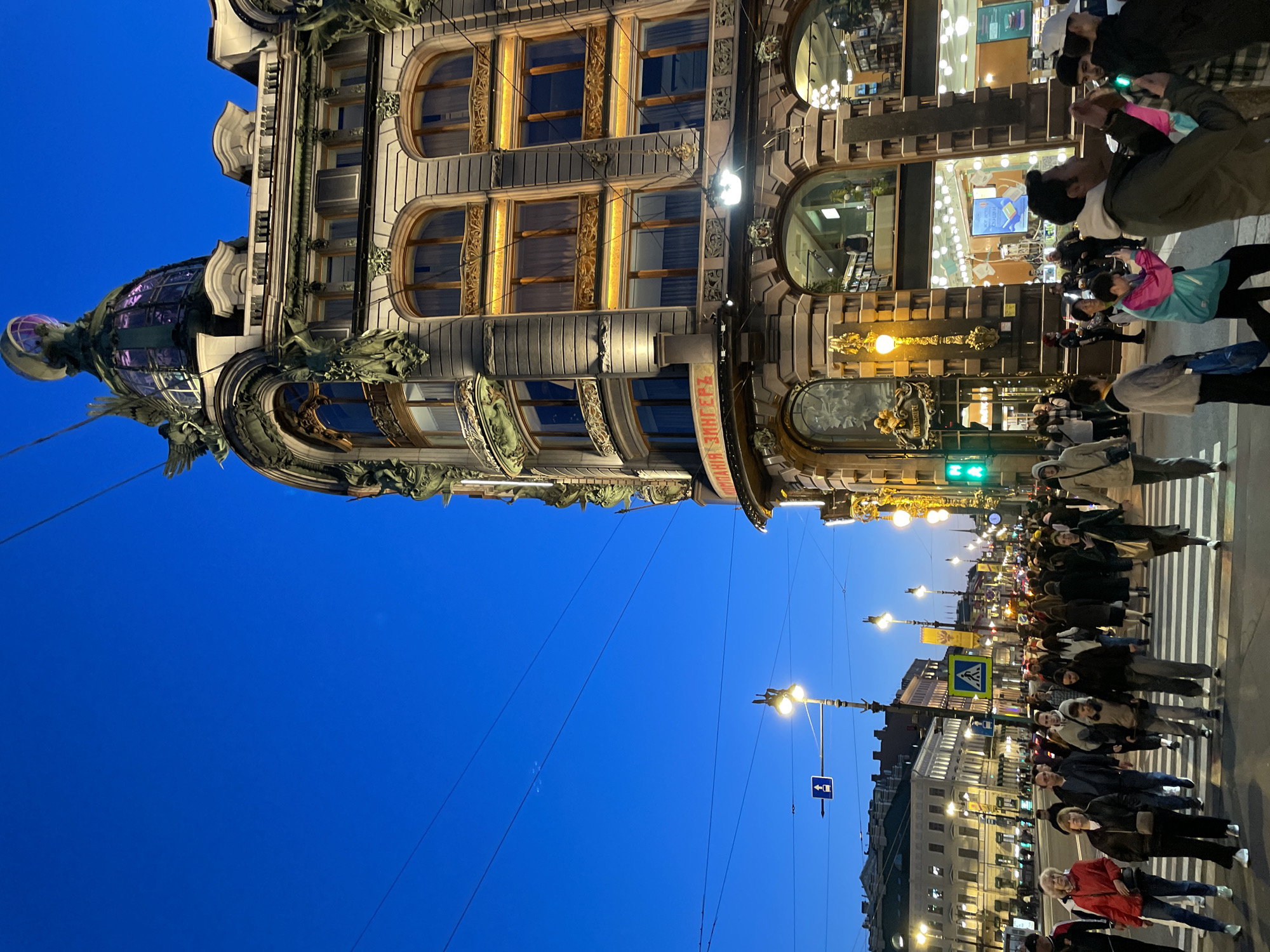 The image presents an evening street scene in Saint Petersburg, Russia, dominated by the iconic Singer House (Dom Knigi). The sky is a deep, dark blue, indicating dusk or night, with the city illuminated by artificial lights.
On the left, the magnificent Singer House stands out with its ornate Art Nouveau architecture. The building features multiple stories with large, arched windows and intricate decorative elements, including sculptures, reliefs, and stylized columns. A striking glass dome or cupola, glowing with a purple hue, crowns the top-left section of the building. Warm, inviting light emanates from its numerous windows, highlighting the architectural details. Along a curved facade section, the text "КОМПАНИЯ ЗИНГЕРЪ" (Singer Company) is visible.
Below and to the right of this grand structure, a bustling street is filled with people. Many individuals are seen walking and crossing a clearly marked pedestrian crossing. They are dressed in various jackets and coats, suggesting cool evening temperatures. Some people are looking up at the Singer House, possibly admiring its beauty or taking photos, while others are engaging with their phones or simply moving along the street.
Streetlights cast bright pools of light on the pavement and illuminate the crowd. A green traffic light is visible near the building, and several street signs, including a blue pedestrian crossing sign and a directional arrow, can be discerned. Buildings with illuminated windows line the street into the distance on the right, contributing to the vibrant urban ambiance. The scene captures a lively, cultural evening in the city.