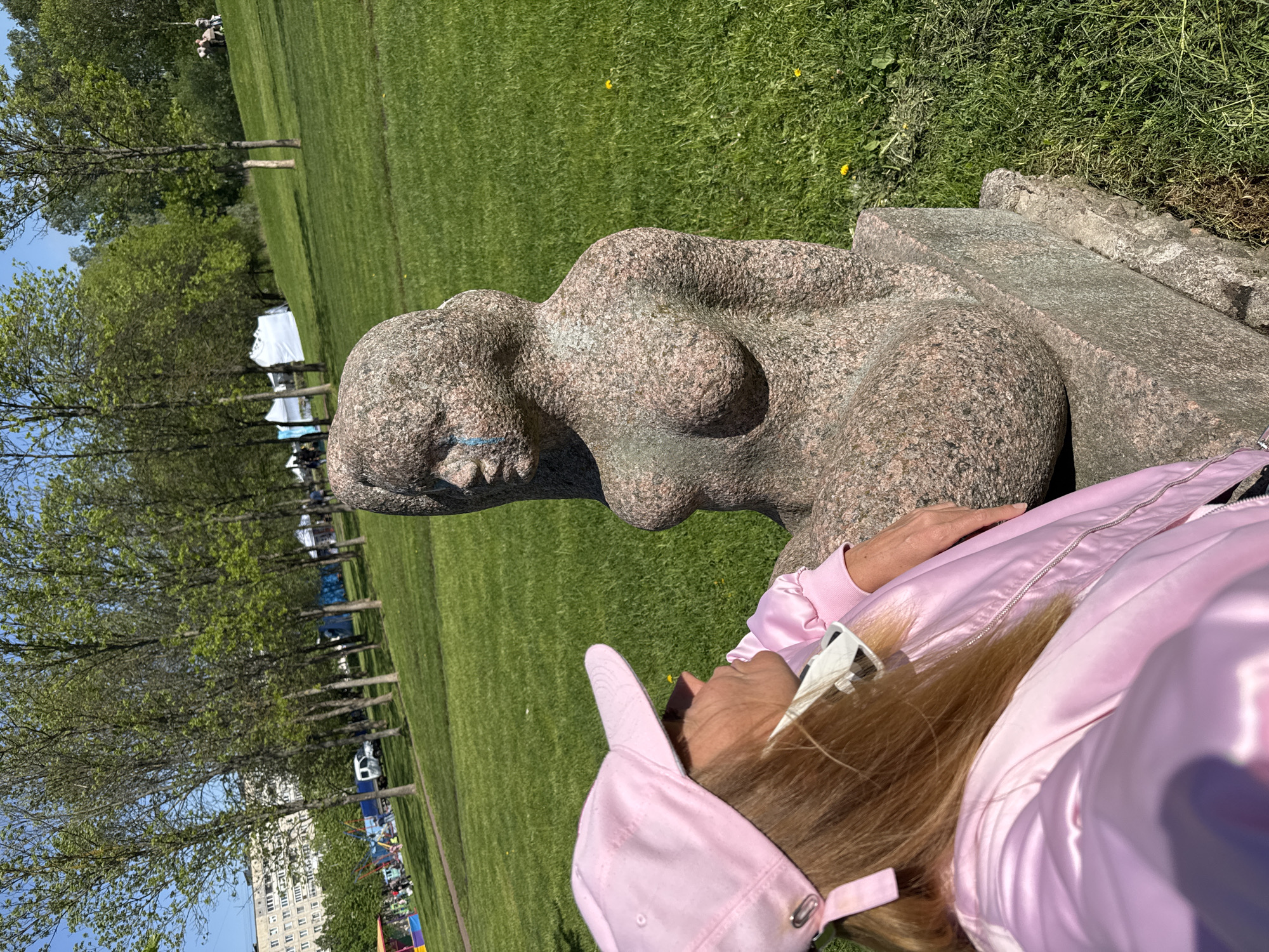 The image shows a woman wearing a pink cap and jacket, resting her head on a stone sculpture of a reclining female figure. The sculpture is weathered and gray, with blue paint markings on the mouth. In the background, there is a grassy park with trees, tents, and a building. The location is likely the park near the Krestovsky Island metro station in Saint Petersburg, Russia.