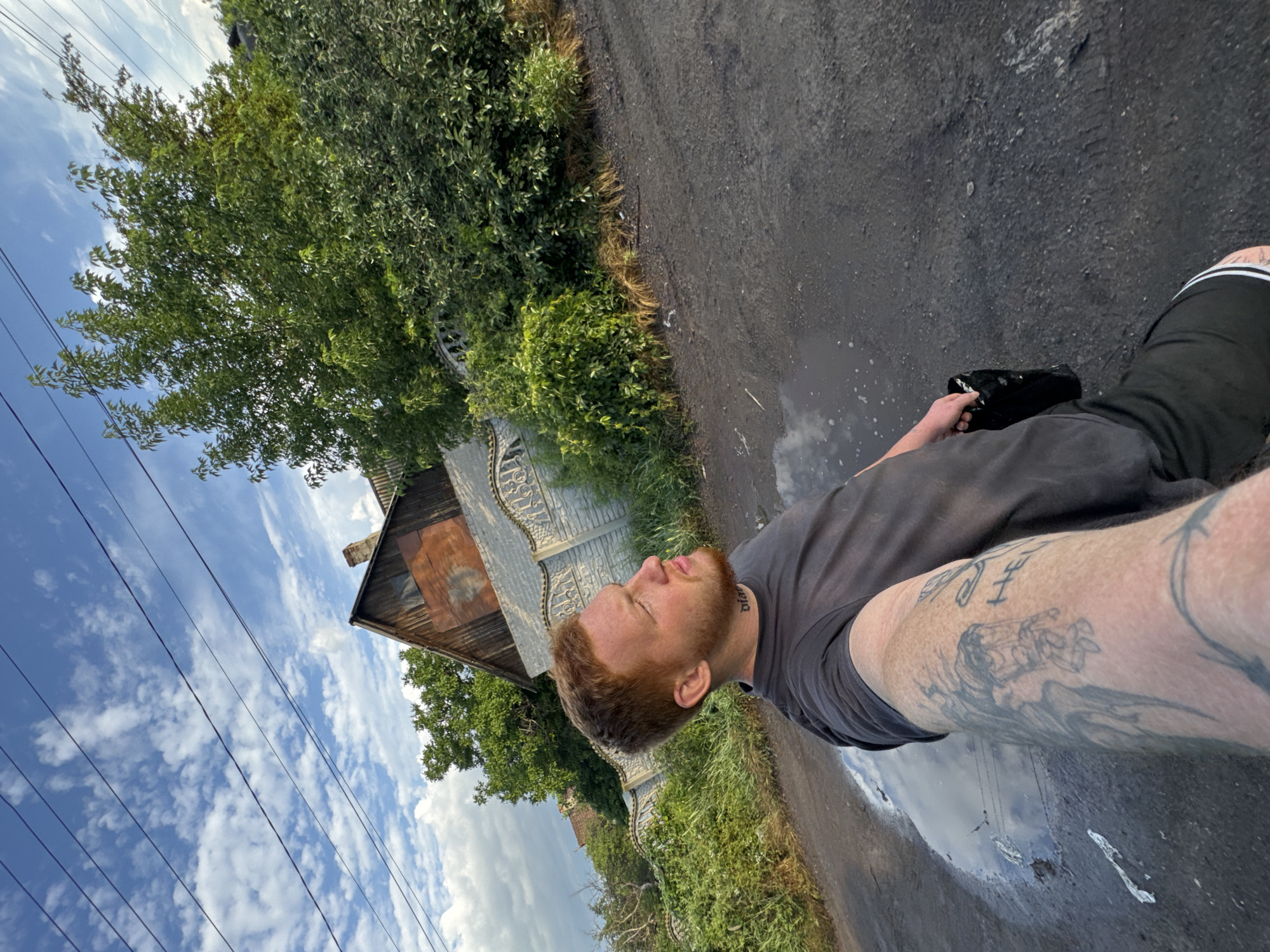 A young Caucasian man with red hair and a beard stands on a dirt road with a large puddle reflecting the sky and clouds. He is wearing a dark gray t-shirt and black shorts with white stripes. The man has visible tattoos on his left arm and neck. He is holding a black plastic bag in his right hand. In the background, there is a fence made of concrete blocks with ornate metal inserts, a wooden house with a rusty metal roof, and dense green trees and bushes. Power lines are visible against a blue sky with scattered white clouds. The scene appears to be in a rural or suburban area with a slightly run-down appearance.