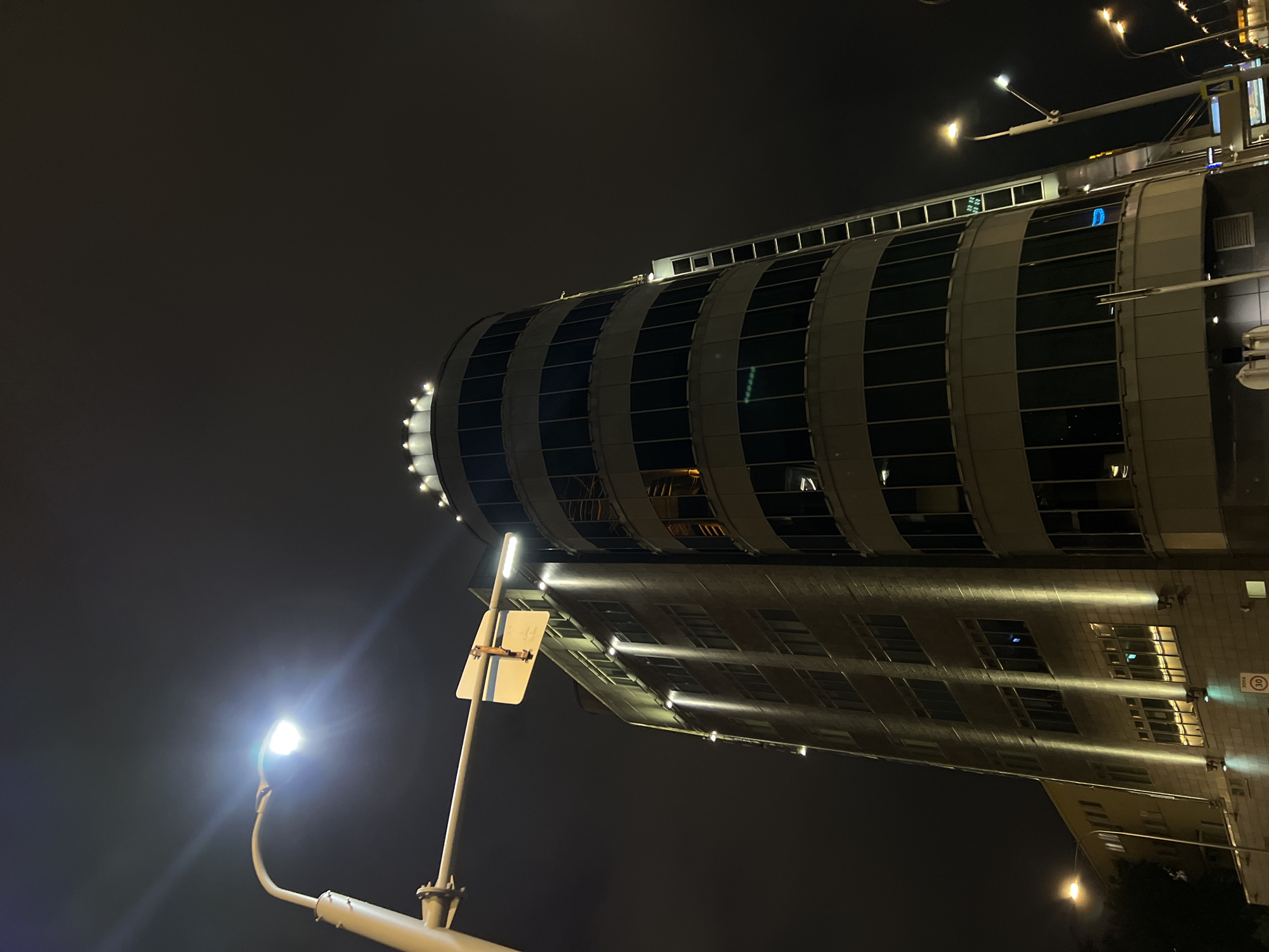 The image depicts a cylindrical building in Moscow, Russia at night. The building features alternating bands of glass and gray material, with lights along the top edge. The building's facade reflects light. A street light and street sign are visible in the foreground. The background is a dark, overcast sky.