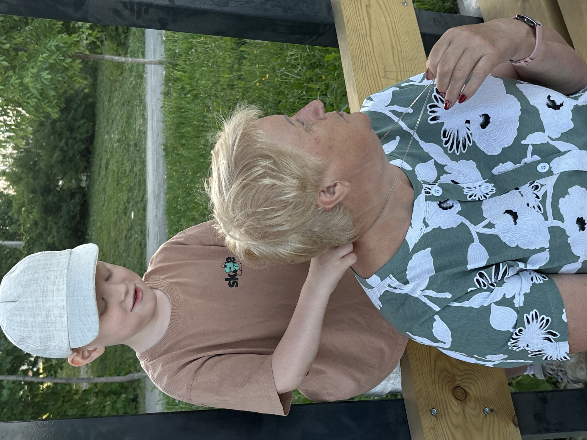 A child with a white hat and a light-brown shirt is facing left, with their left hand on the back of a person with short blonde hair. The person is lying on a wooden bench, wearing a green top with large white flowers, and has a silver chain around their neck. The scene is outdoors, likely in a park, given the green grass and trees in the background. The location is possibly Izhevsk, Russia, but there are no explicit contextual cues.
