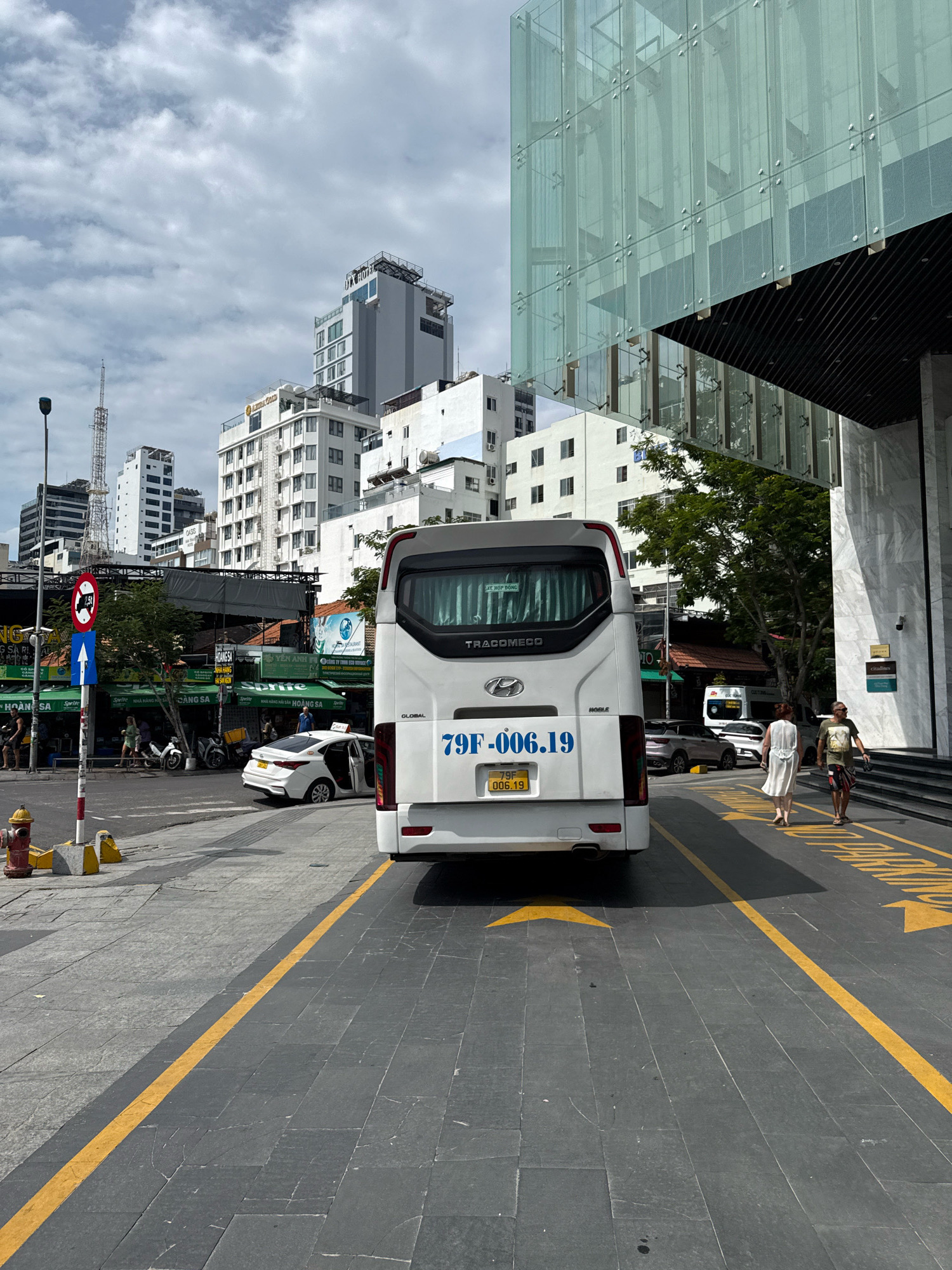 The image shows a street scene in Nha Trang, Vietnam, dominated by a white bus with license plate 79F-006.19. The bus is parked on the right side of the road, partially obscuring a modern building with a glass facade. A white taxi with its doors open is visible to the left of the bus, and a few pedestrians are walking on the sidewalk. The street is paved with dark gray tiles and marked with yellow lines. In the background, several buildings of varying heights, including high-rises, are visible under a cloudy sky. A red fire hydrant is located on the sidewalk on the left side of the image. The overall impression is that of a bustling urban environment.