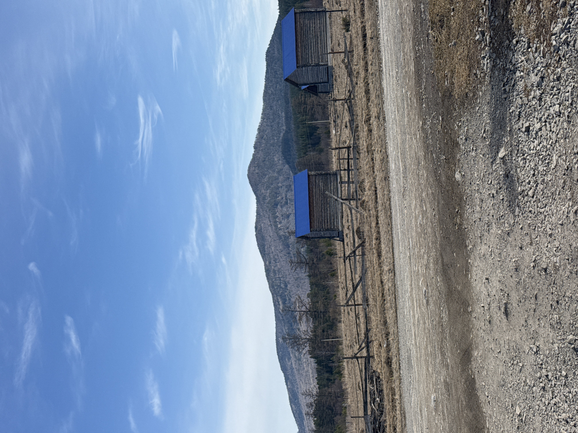 The image shows a rural landscape in the village of Tyulyuk, Russia. Two wooden structures with blue roofs stand parallel to a dirt road. A dilapidated wooden fence runs along the edge of the road, separating it from a field of dry grass. In the background, a mountain rises into a clear blue sky with scattered wispy clouds. The ground is covered with gravel. No people or animals are visible.