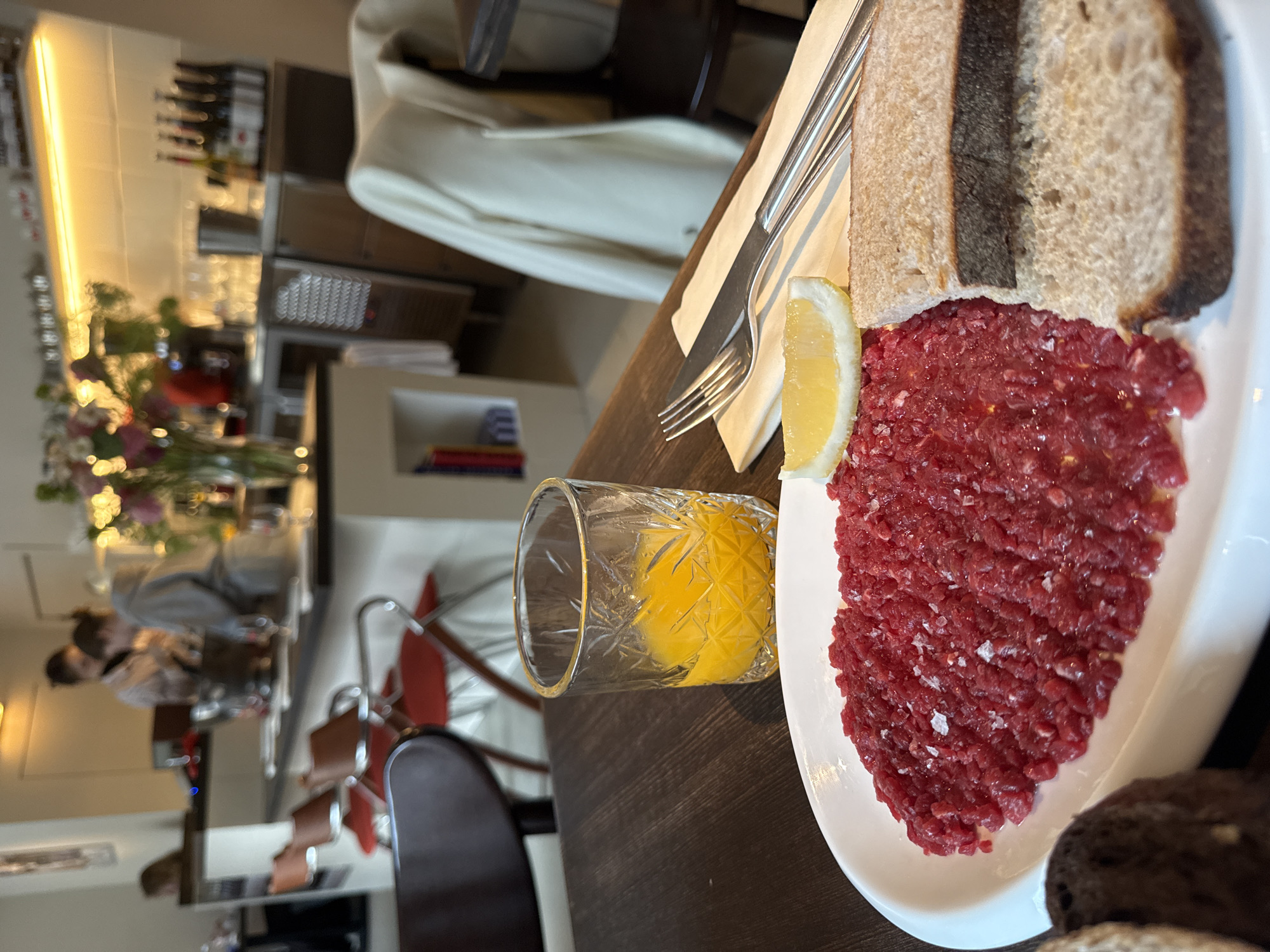 The image shows a plate of steak tartare with slices of dark bread and a lemon wedge, accompanied by a fork and knife. A glass of orange juice is nearby. The scene is set in a restaurant, with a blurred view of other diners and staff in the background. The restaurant appears to be located in Moscow, Russia, based on the style of the interior design, furniture, and the overall ambiance.