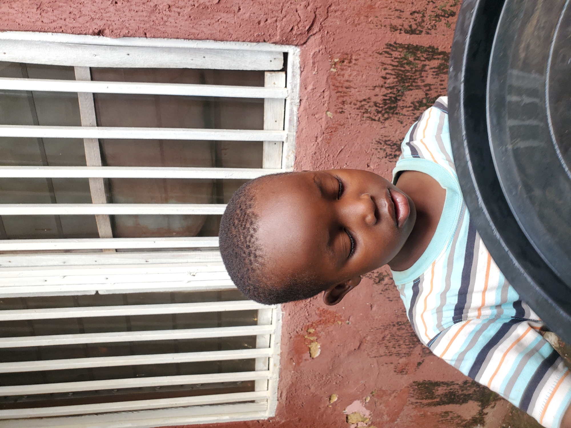 In Jos, Nigeria, a young boy with dark skin and short hair is captured in a medium shot, his eyes closed, suggesting a moment of rest or contemplation. He is wearing a light blue t-shirt with horizontal stripes in white, orange, navy, and light blue. His head is slightly bowed, and his gaze is directed downwards. The boy is positioned in front of a red, textured wall, which appears weathered and shows signs of age, with patches of lighter color and dark organic growth. To the left of the boy, a white-barred window is visible, adding a linear element to the background. The bars are vertical and spaced apart, allowing a glimpse of the interior, which appears to be dimly lit. In the foreground, a dark, round object, possibly a large plastic basin or container, partially obscures the boy's lower body and the lower part of the wall. The overall lighting suggests an outdoor setting, with the light source likely from above and slightly to the right, casting subtle shadows on the boy's face and the wall. The scene is devoid of any other people or apparent activity, focusing solely on the solitary figure of the child and his immediate surroundings.